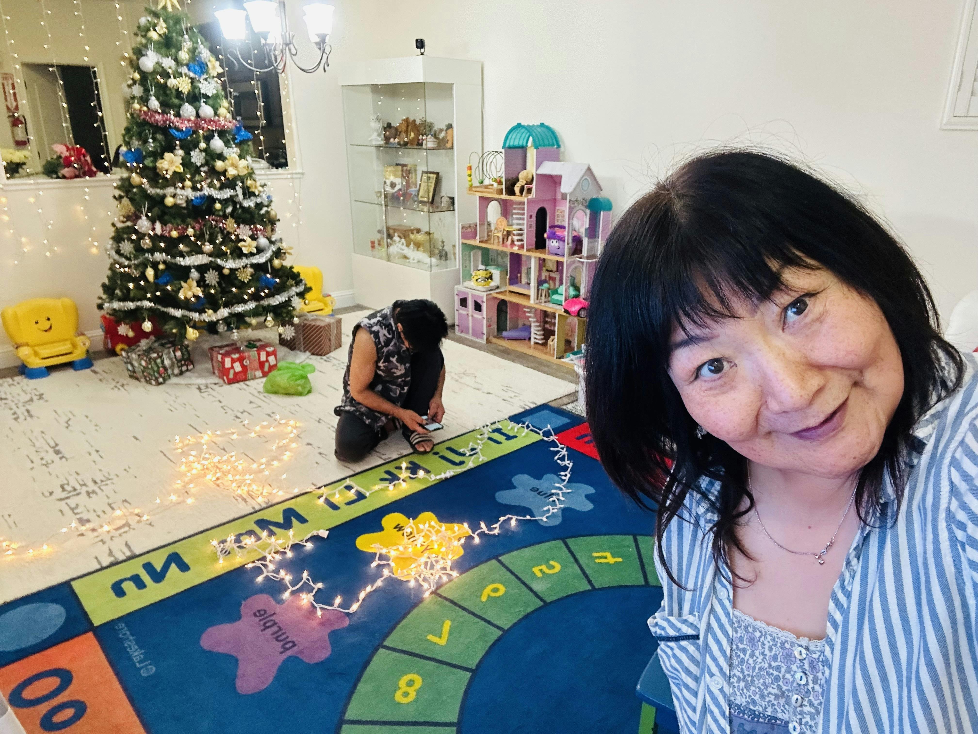 Person taking a selfie with someone arranging string lights on a playroom rug
