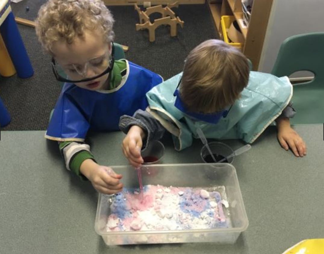 Two children in safety goggles and smocks playing with colored powder in a tray