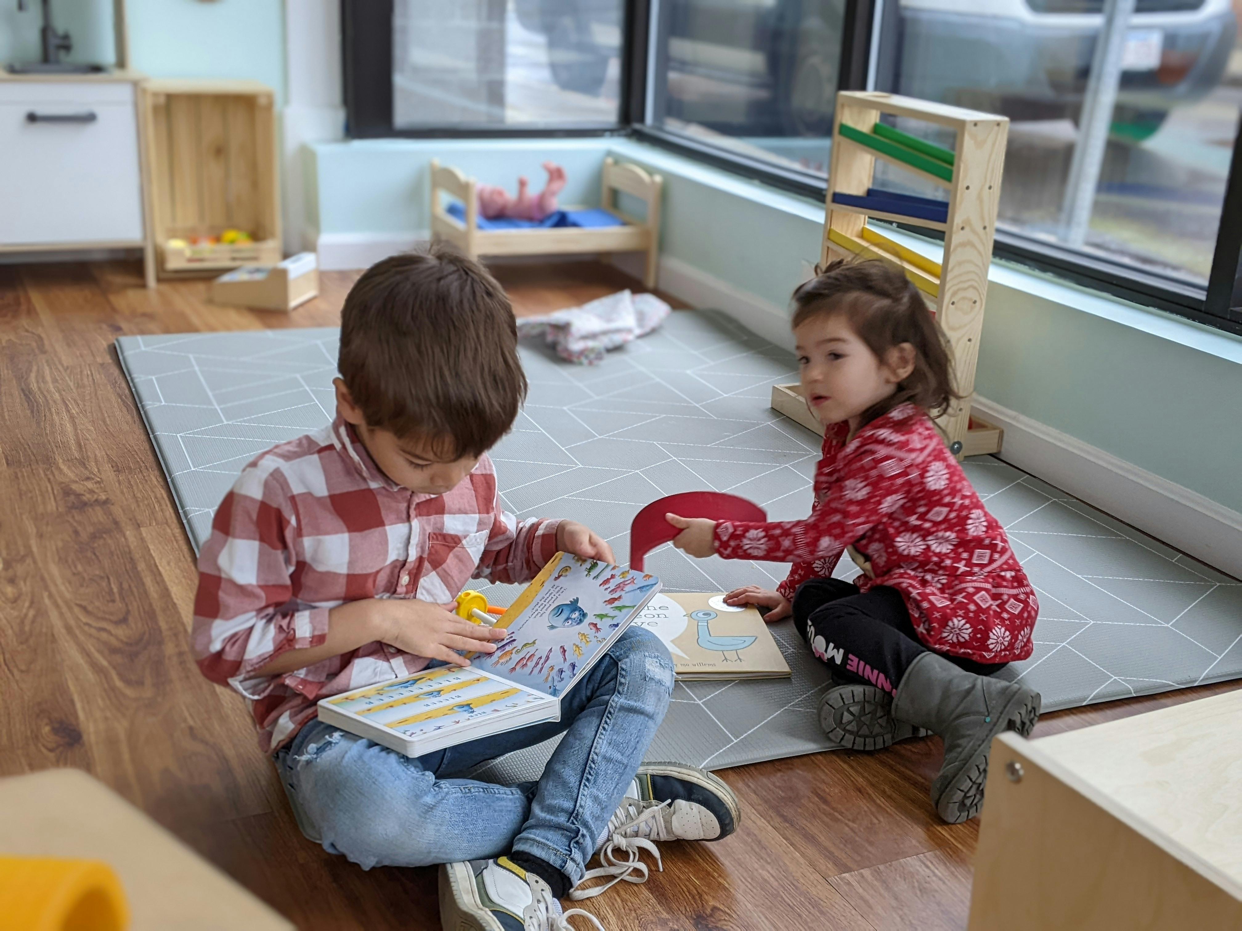 Two children sitting on the floor, one reading a book and the other pointing