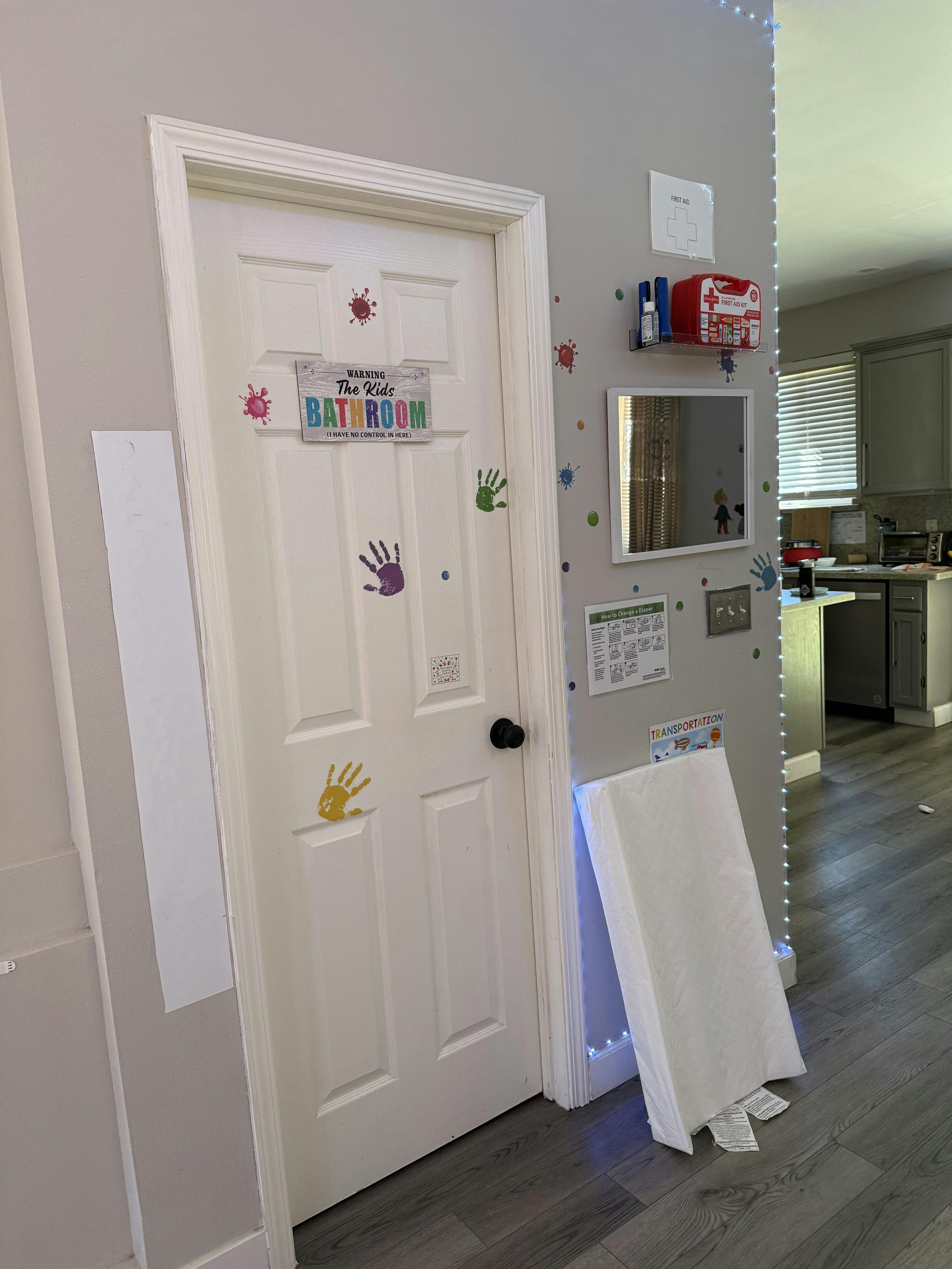 Door labeled The Kids Bathroom with handprints and a changing pad on the wall