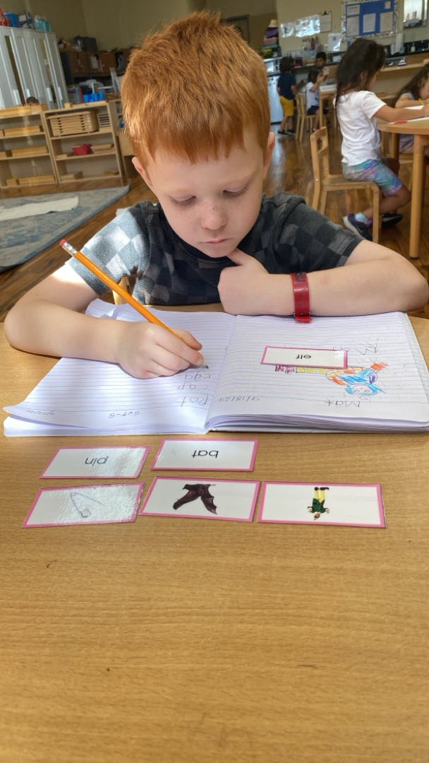 Child writing in a notebook at a table with word cards