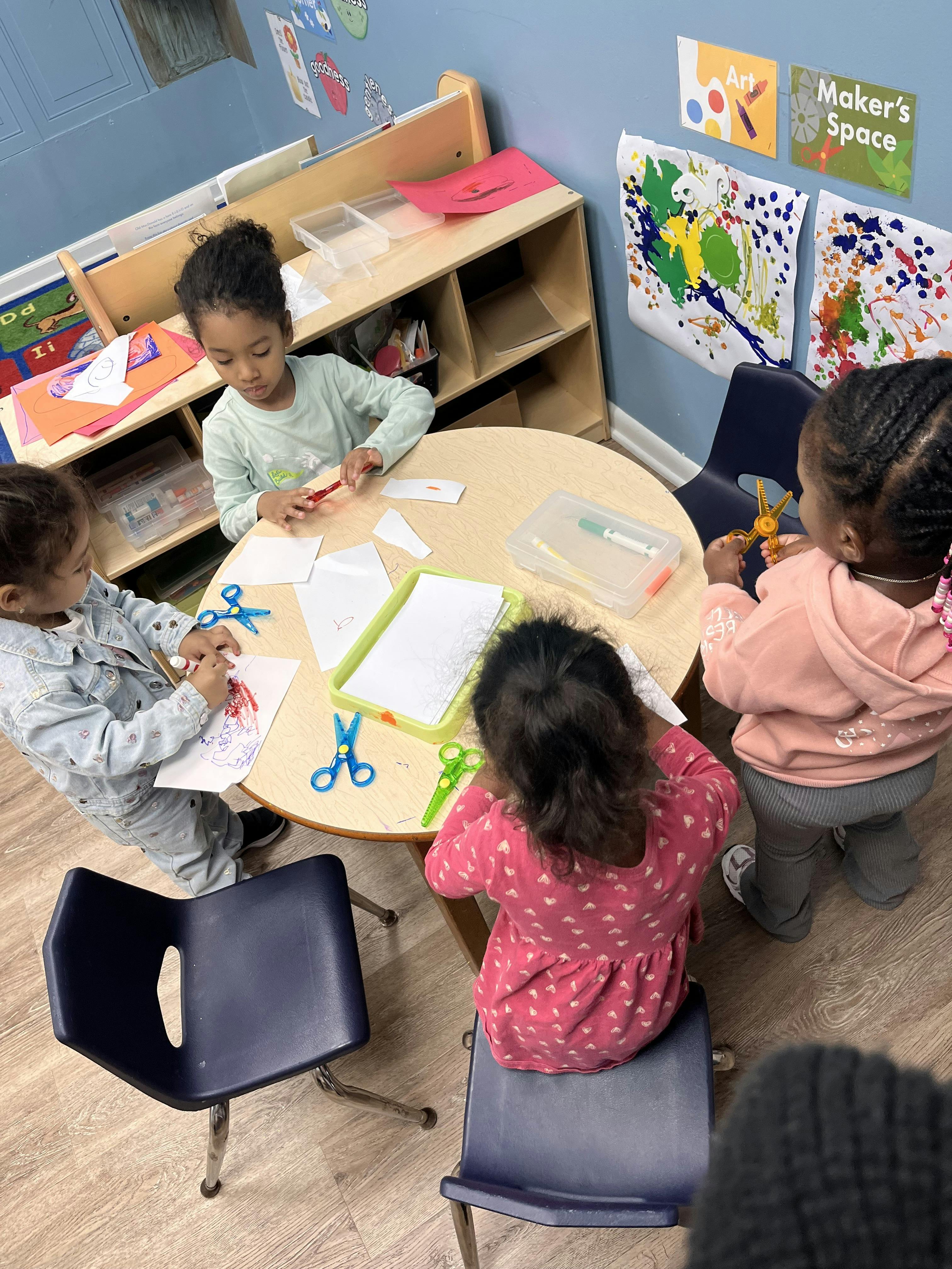 Four children at a round table doing crafts with paper and scissors in a classroom