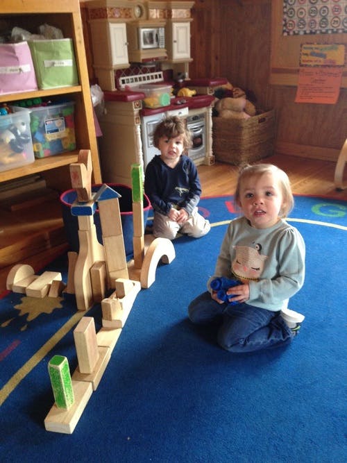 Two children kneeling on a blue rug next to wooden blocks