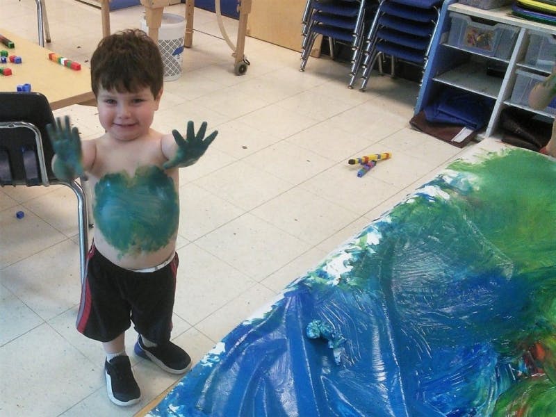 Child with green paint on hands and torso in a classroom