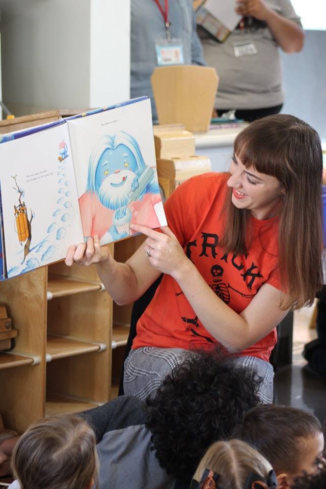 Person reading a book to children, pointing at an illustration of a blue character