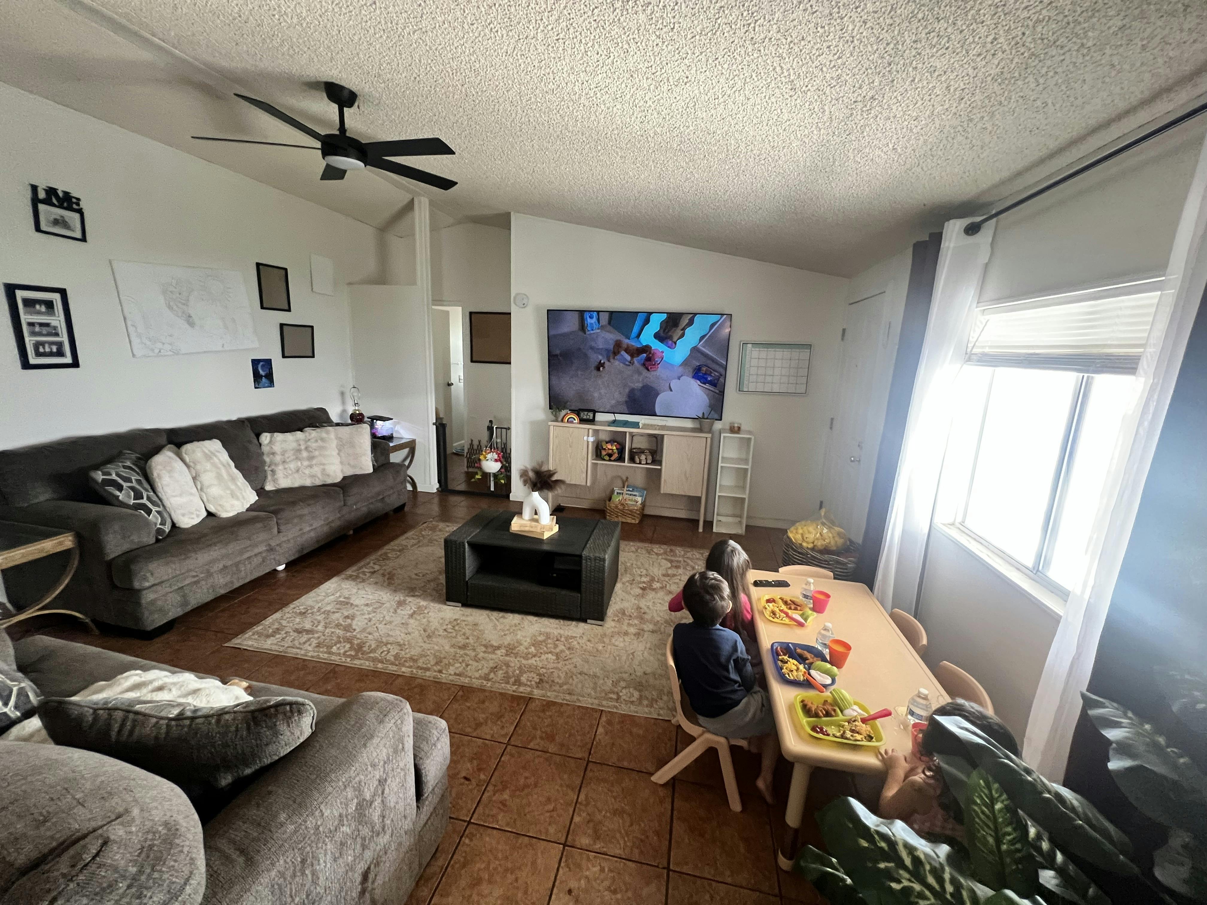 Three children eating lunch at a small table while watching a television