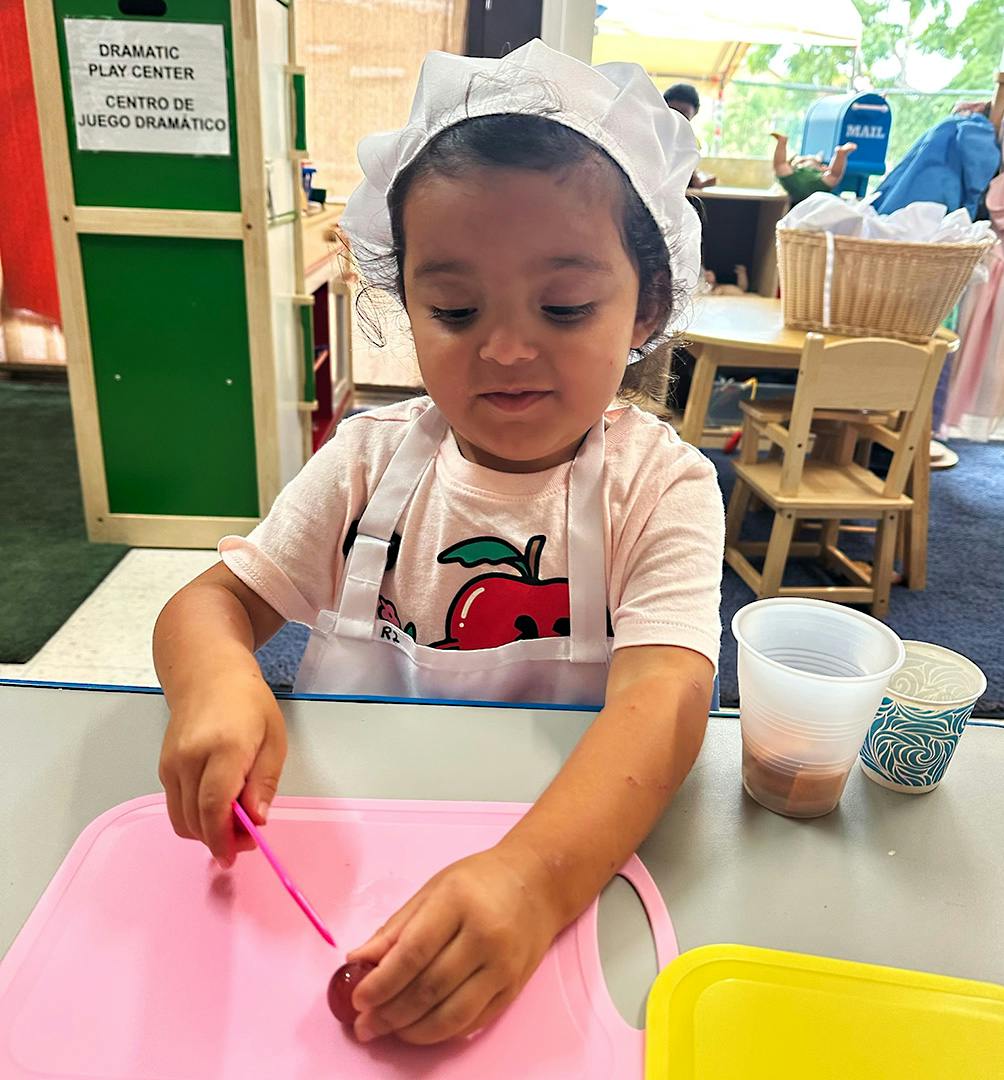Child wearing a chef hat and apron pretending to cut fruit on a cutting board
