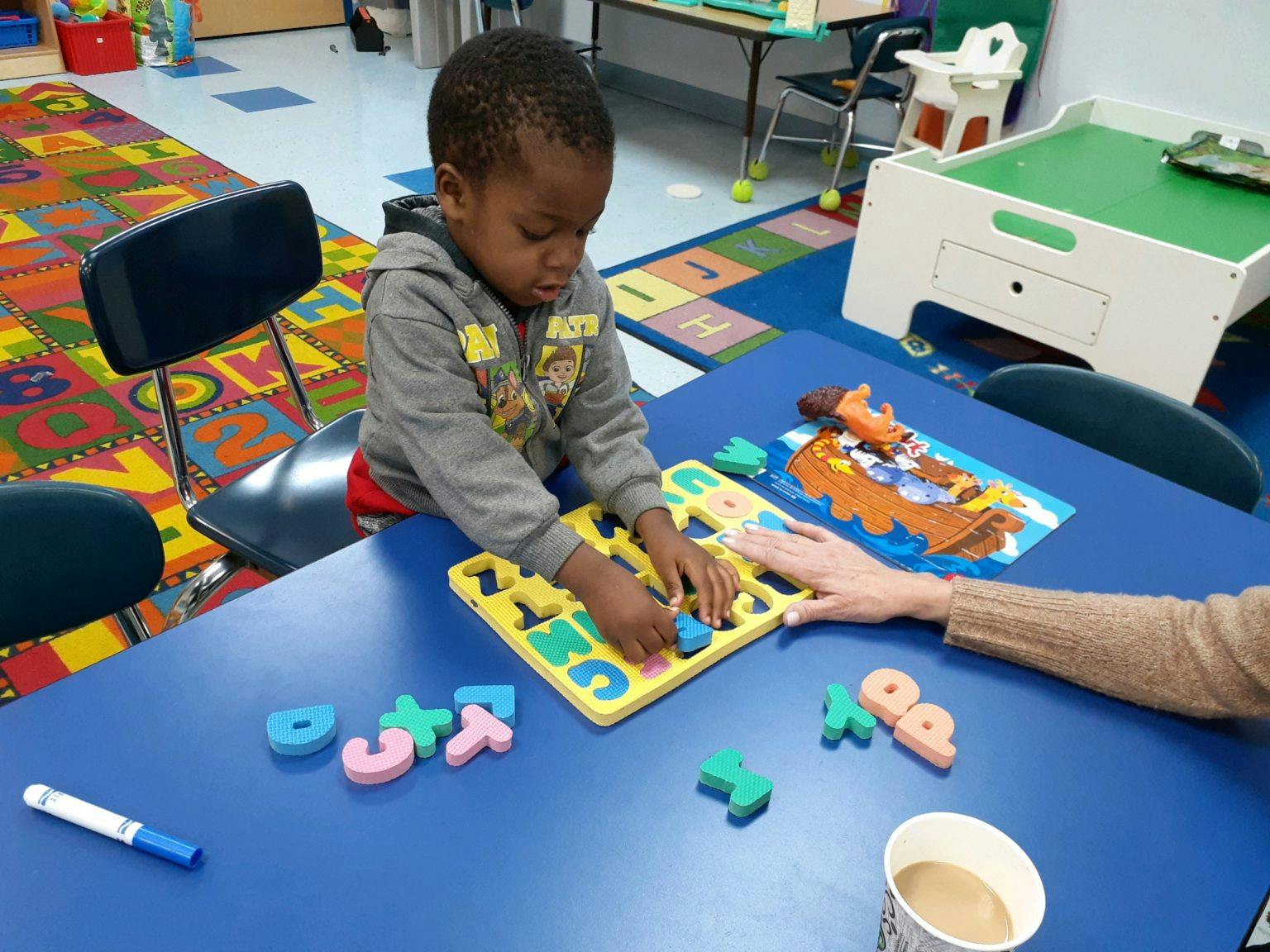 Child playing with a letter puzzle on a blue table with an adult's hand nearby