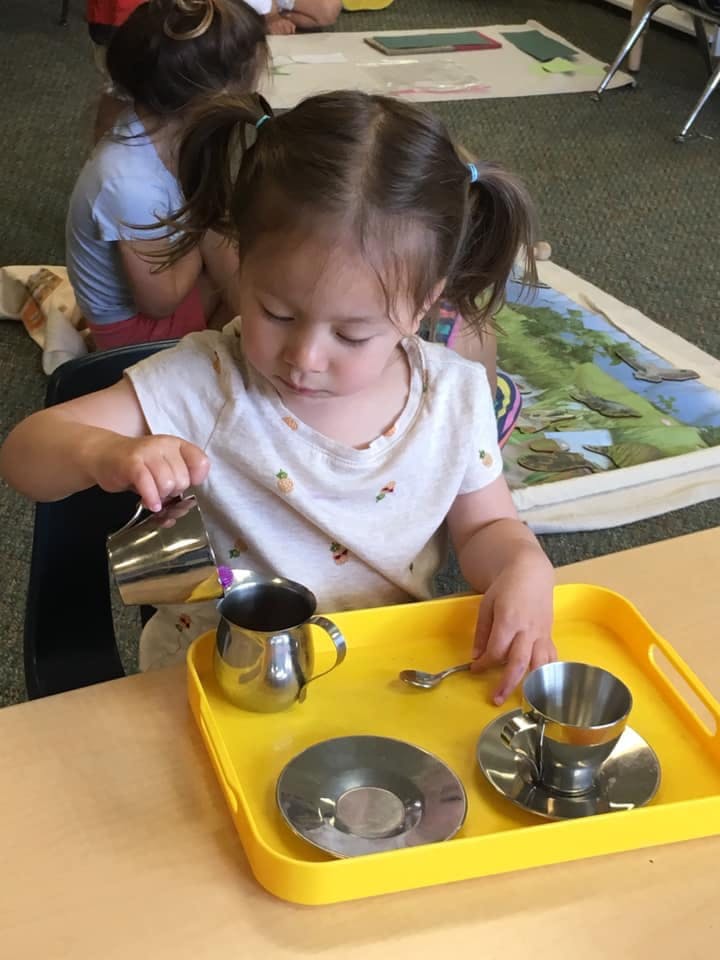 Child pouring from a small metal pitcher into a metal cup on a yellow tray