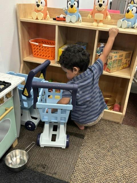 Child playing with a toy shopping cart near a shelf of toys