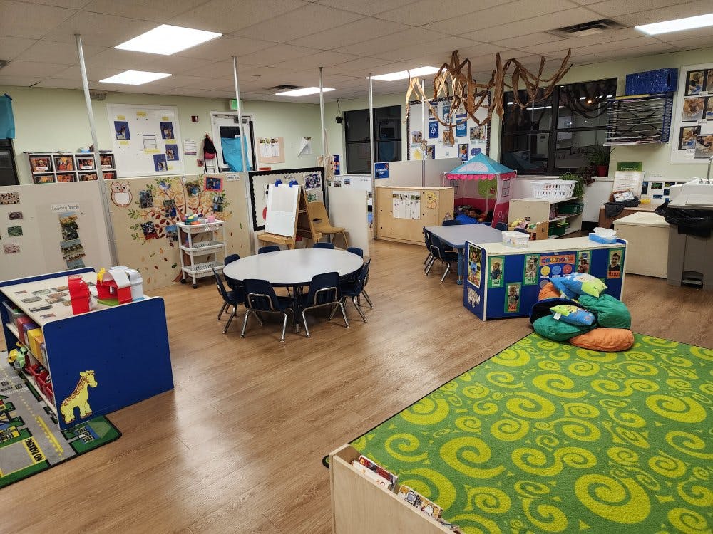 Empty classroom with small tables and chairs, play areas, and a green patterned rug