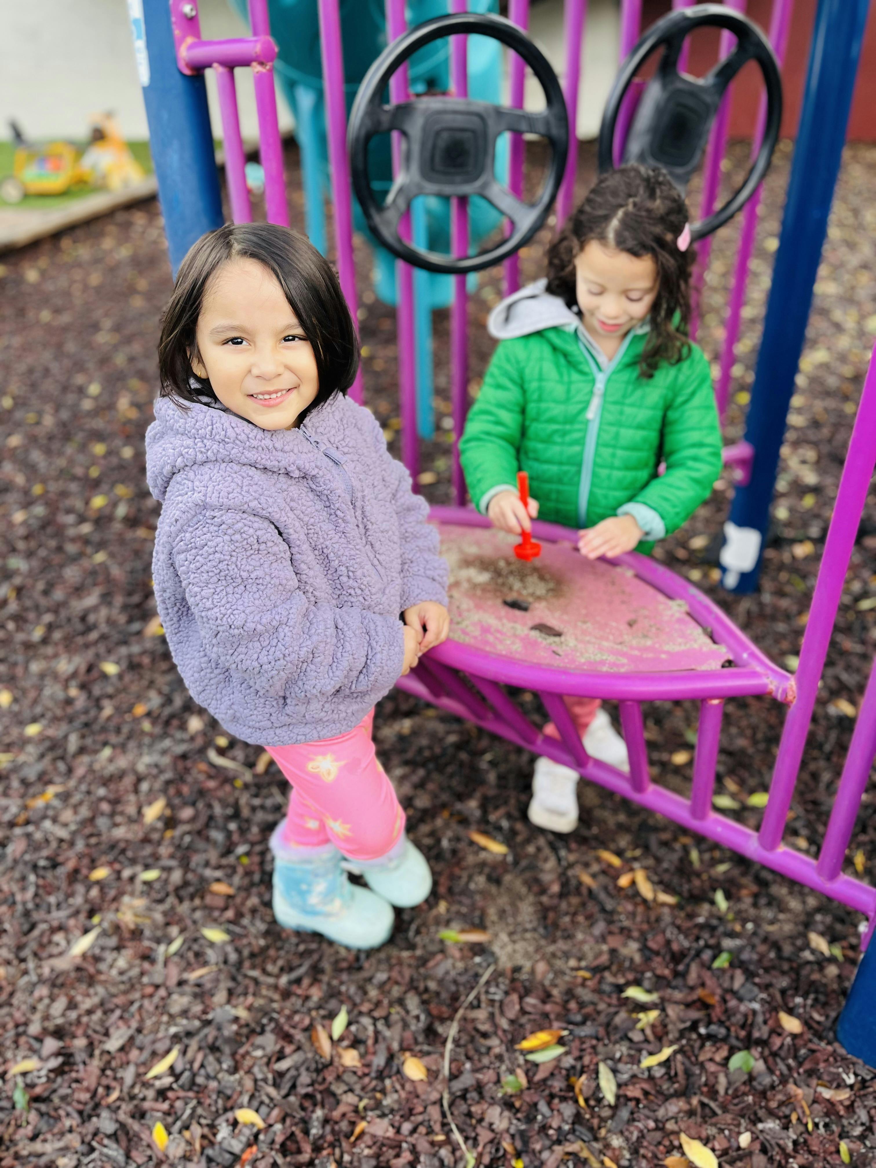 Child smiling at camera while another child plays in a sandbox at a playground