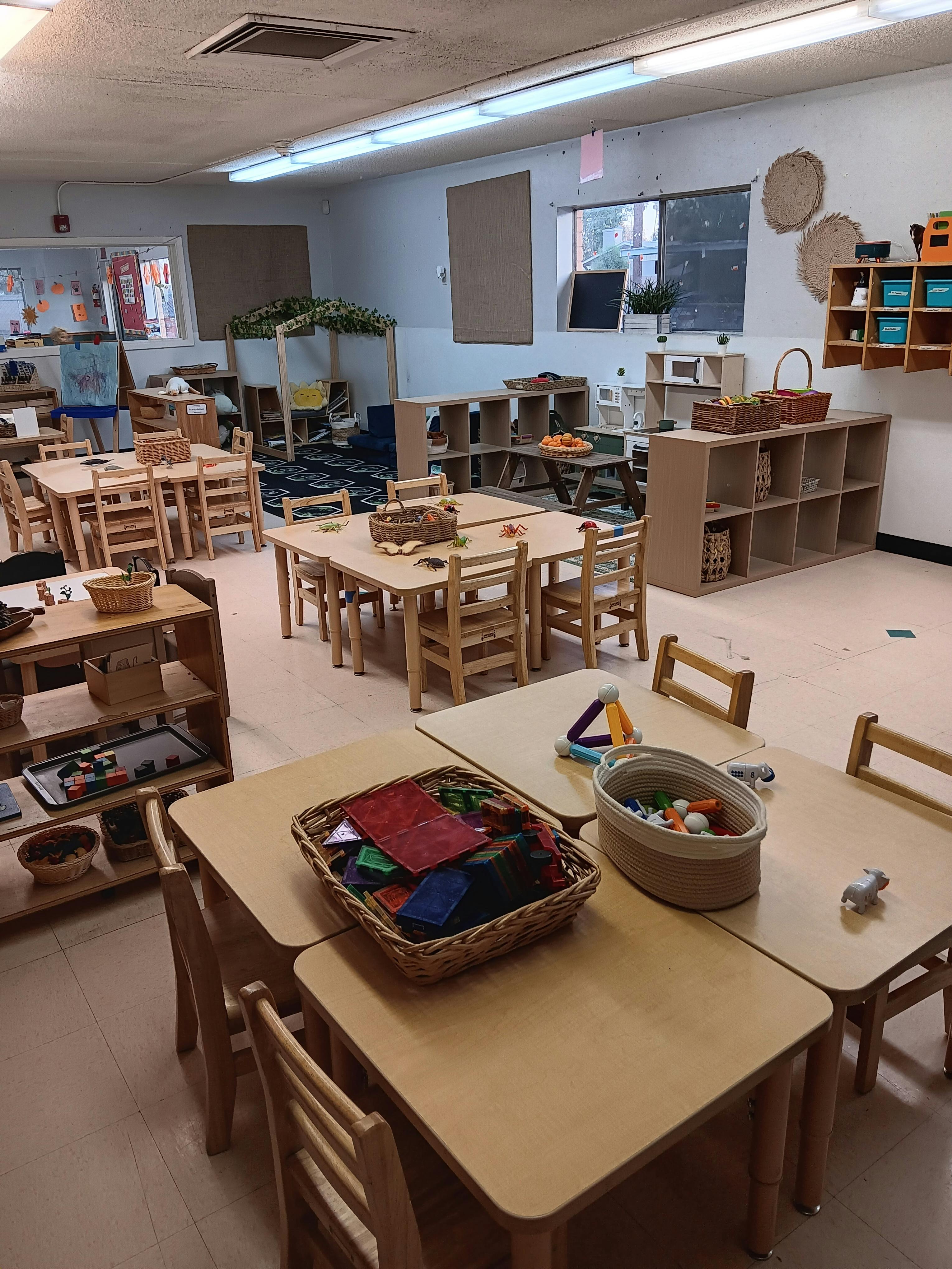Classroom with small wooden tables and chairs, shelves with learning materials