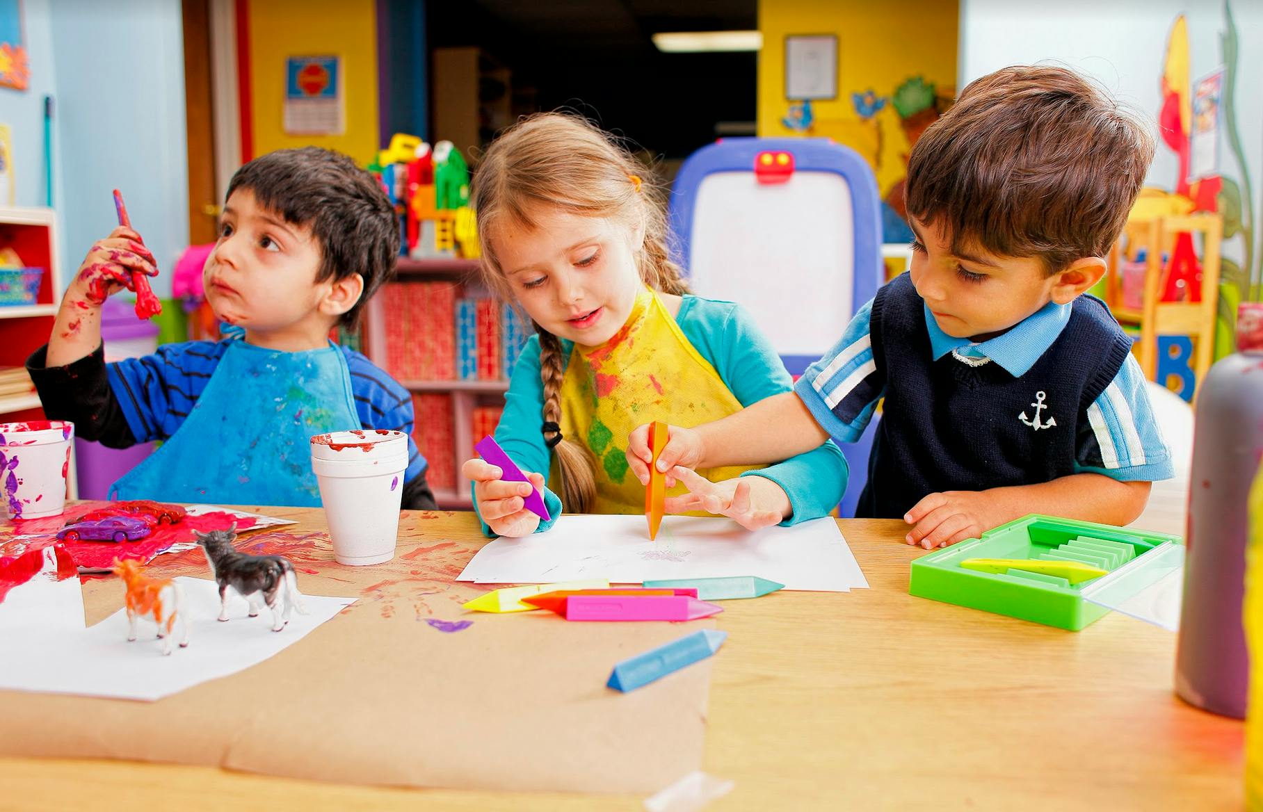 Three children painting and drawing at a table in a classroom