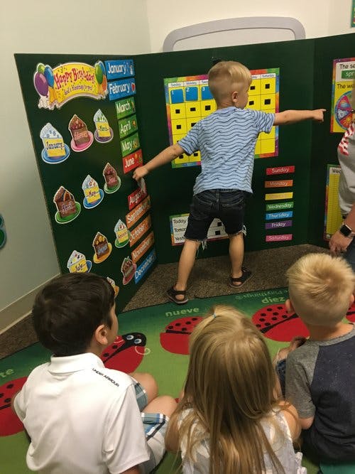 Child standing and pointing at a calendar board while other children sit and watch