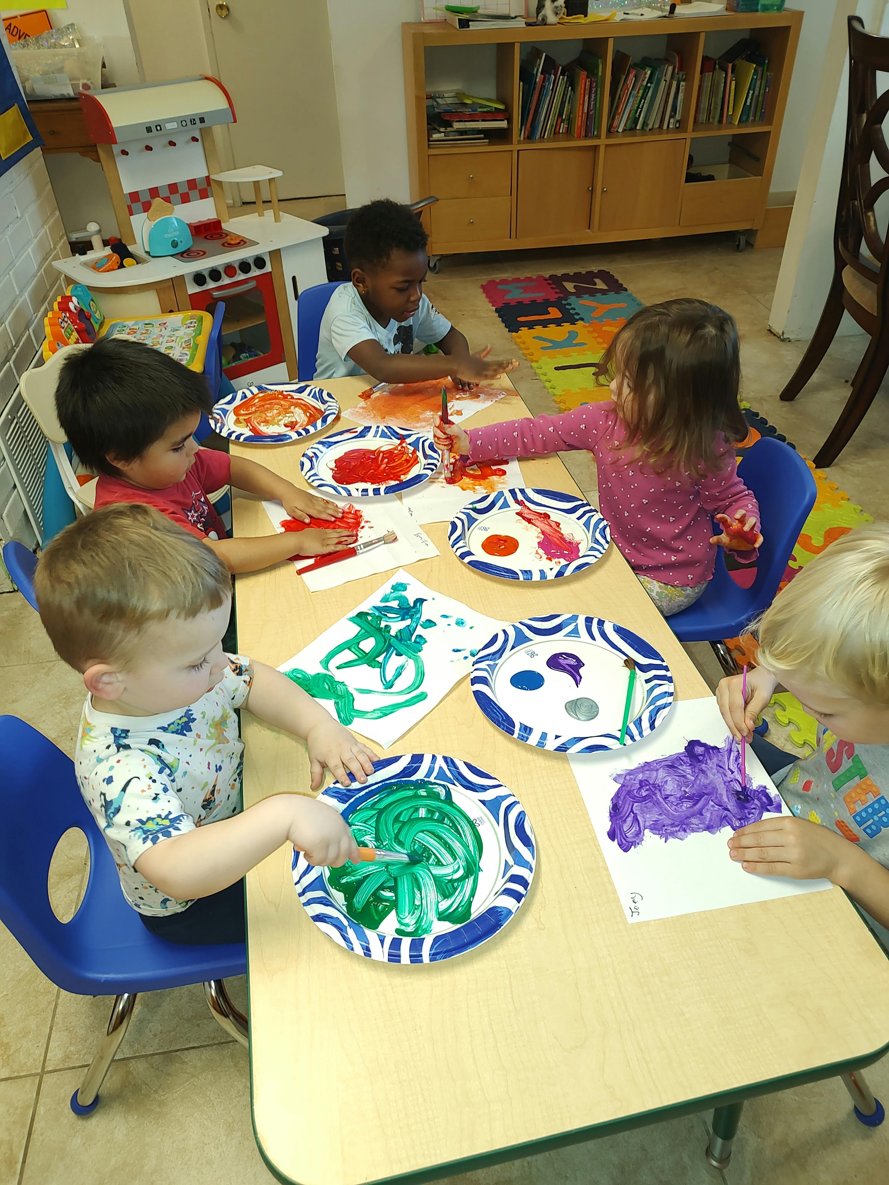 Several children painting with brushes and fingers at a classroom table