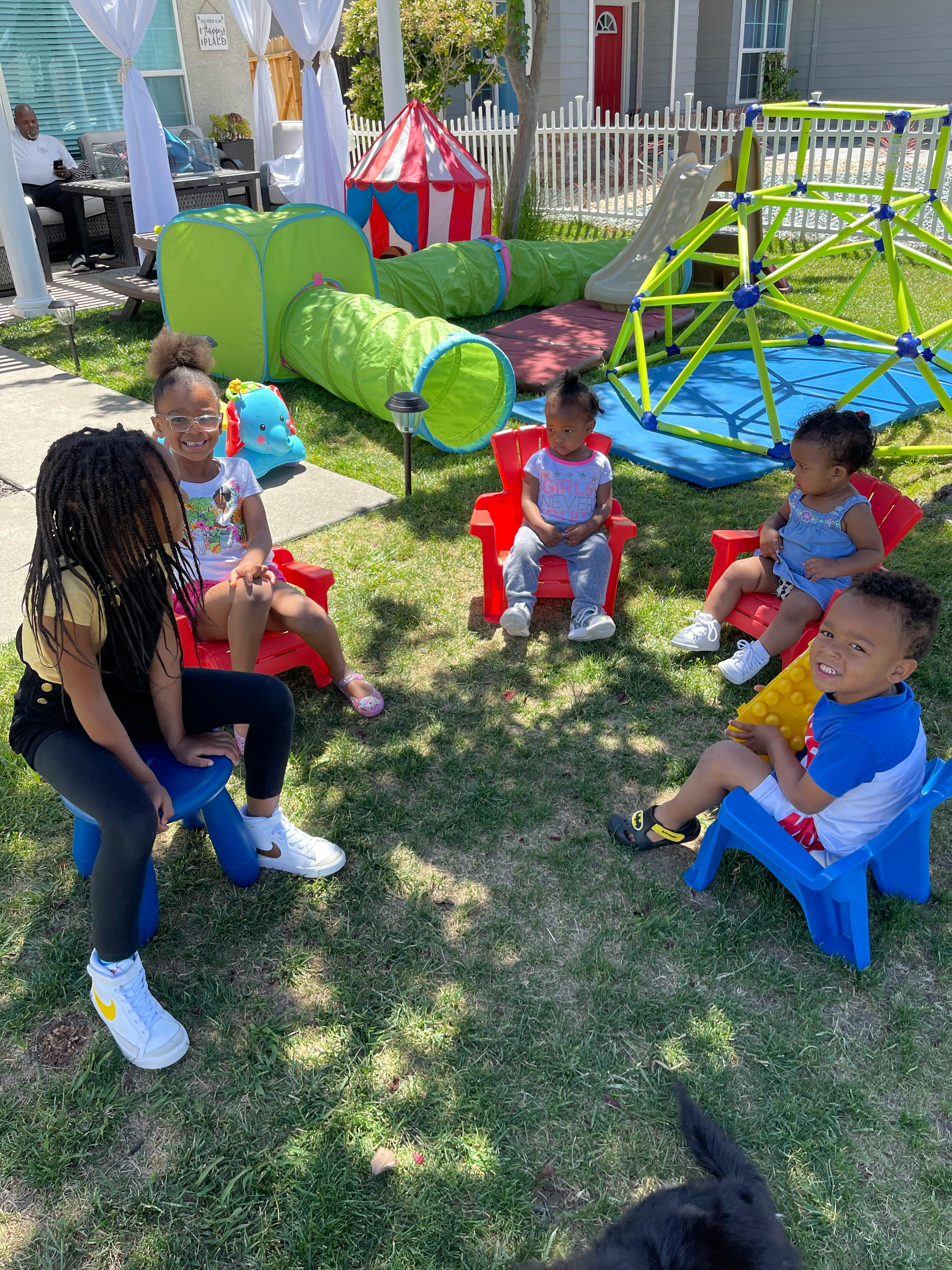 Children sitting in small chairs on grass with play equipment in background