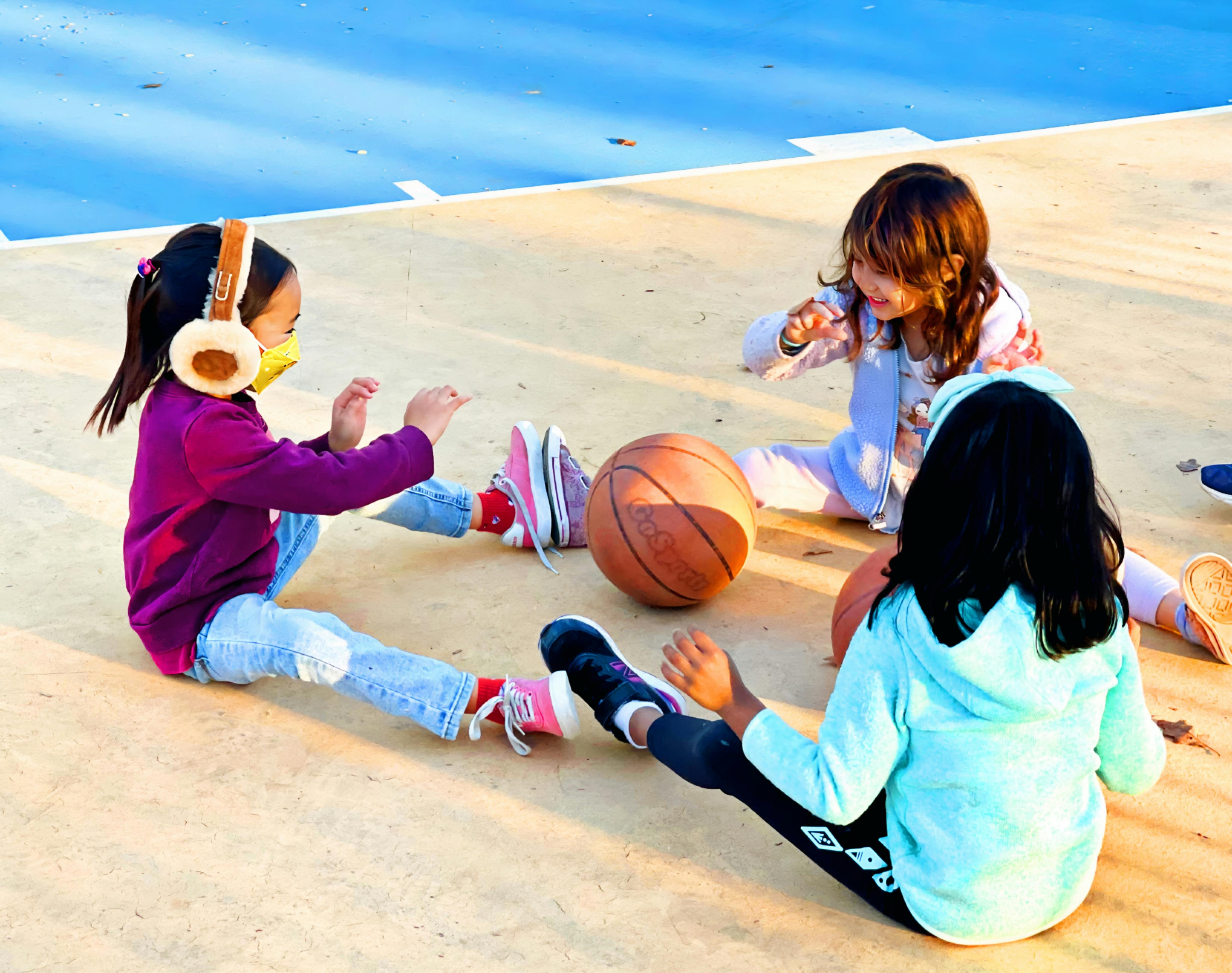 Three children sitting on a court with a basketball