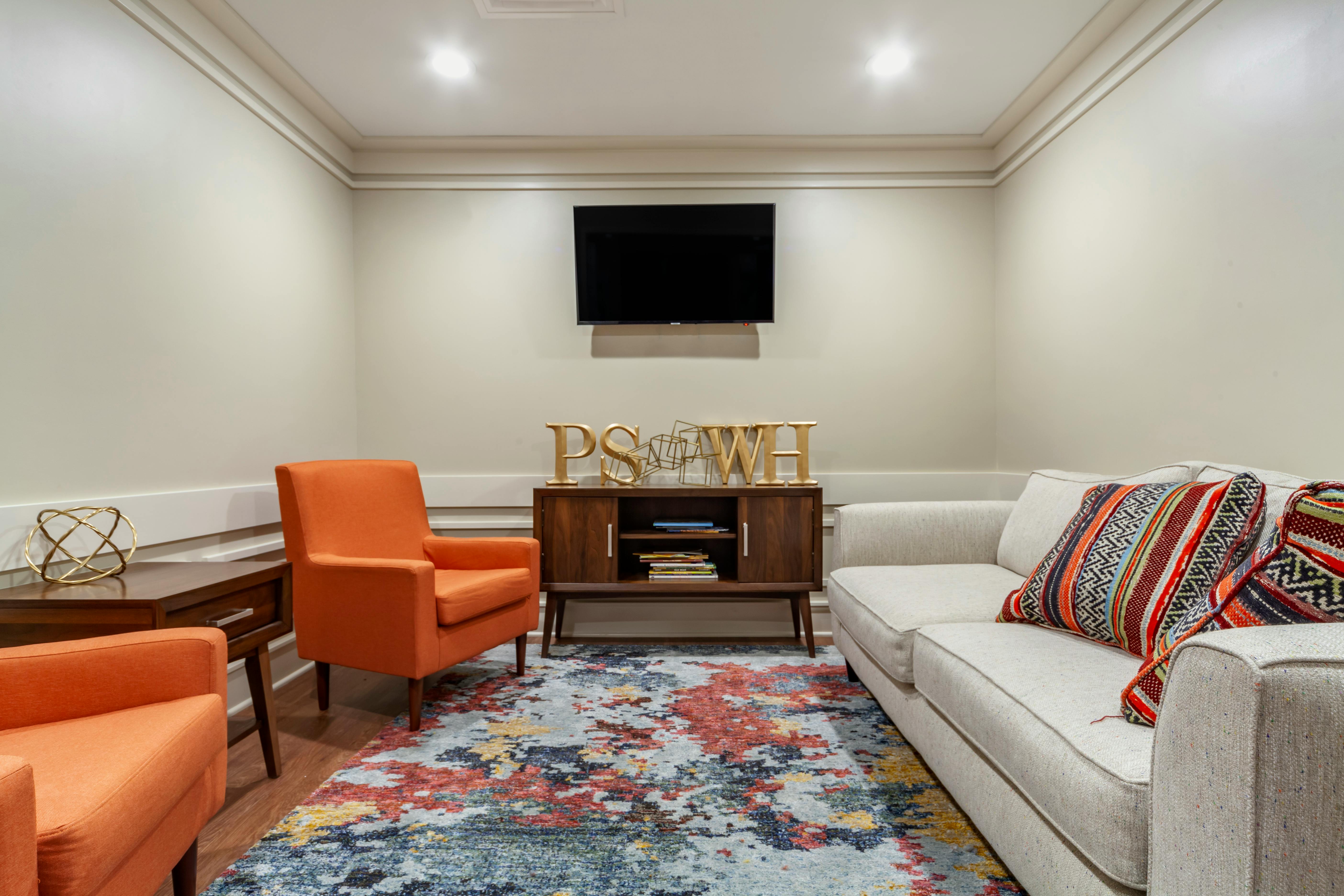Living room with orange chairs, light-colored couch, colorful rug, and a TV