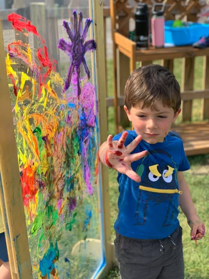 Child with paint on hand next to a clear easel with colorful paint