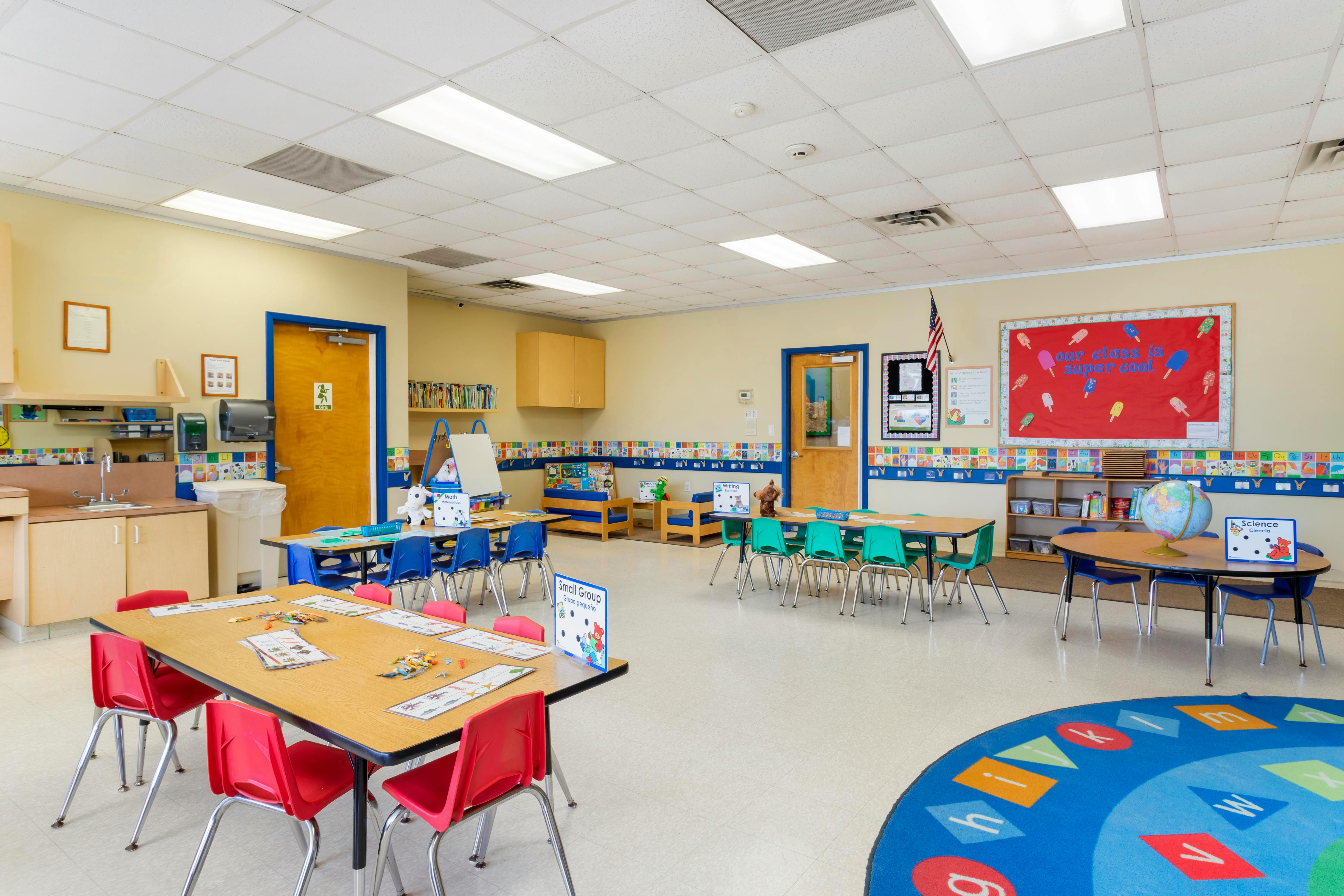 Empty classroom with small tables and chairs, a sink, and a colorful alphabet rug