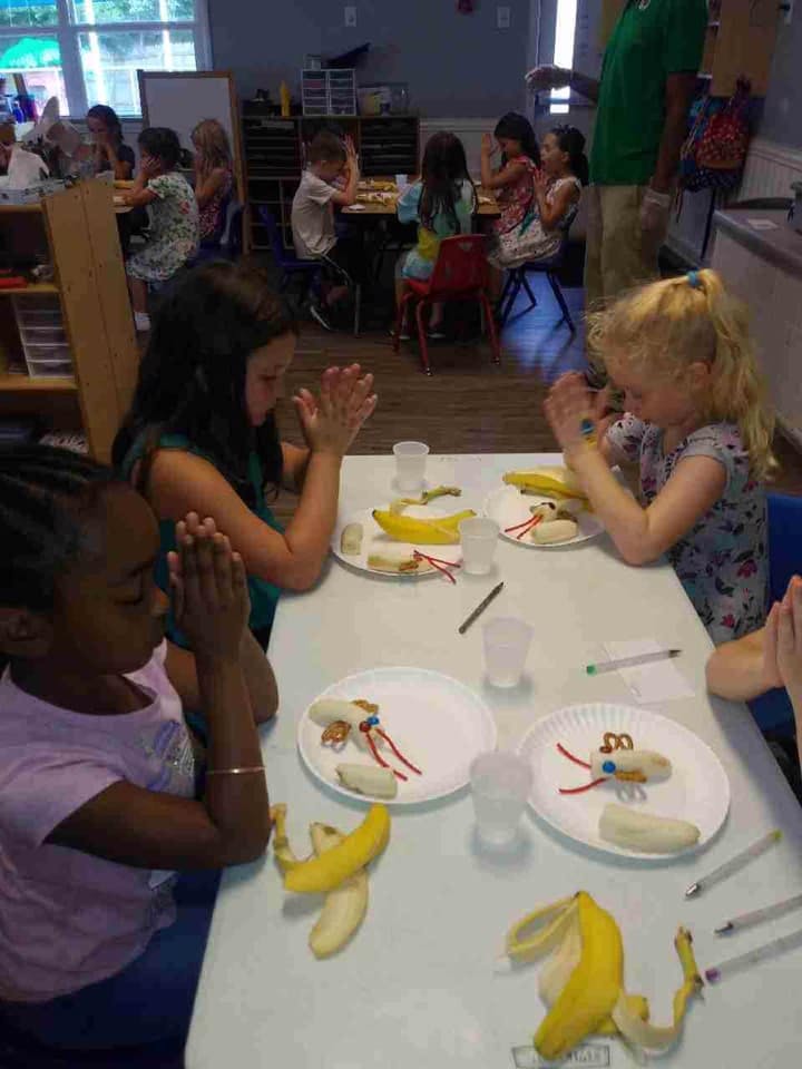 Children and adults sitting at tables with hands together, food on plates