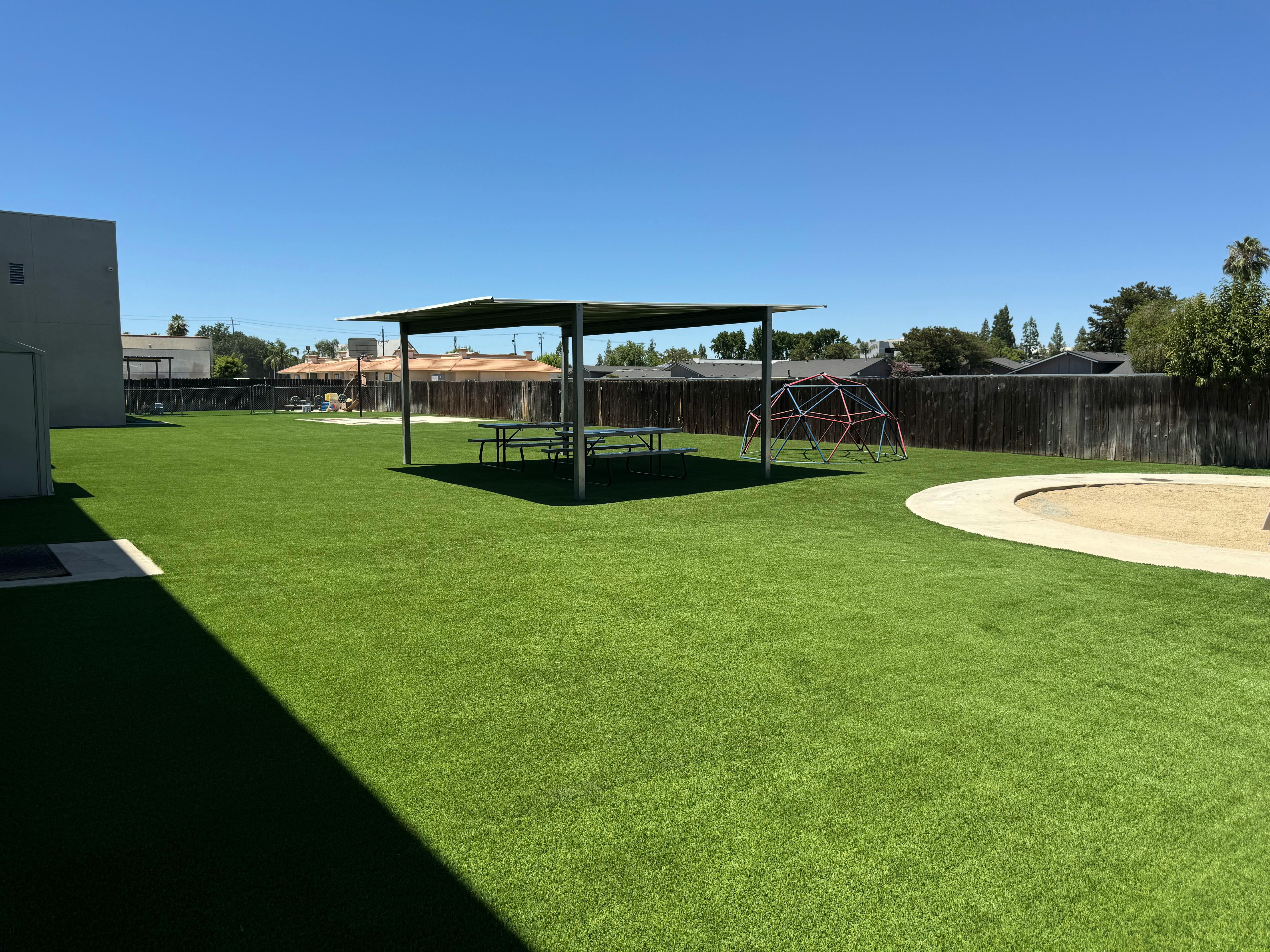 Outdoor play area with artificial turf, covered picnic tables, and sand pit