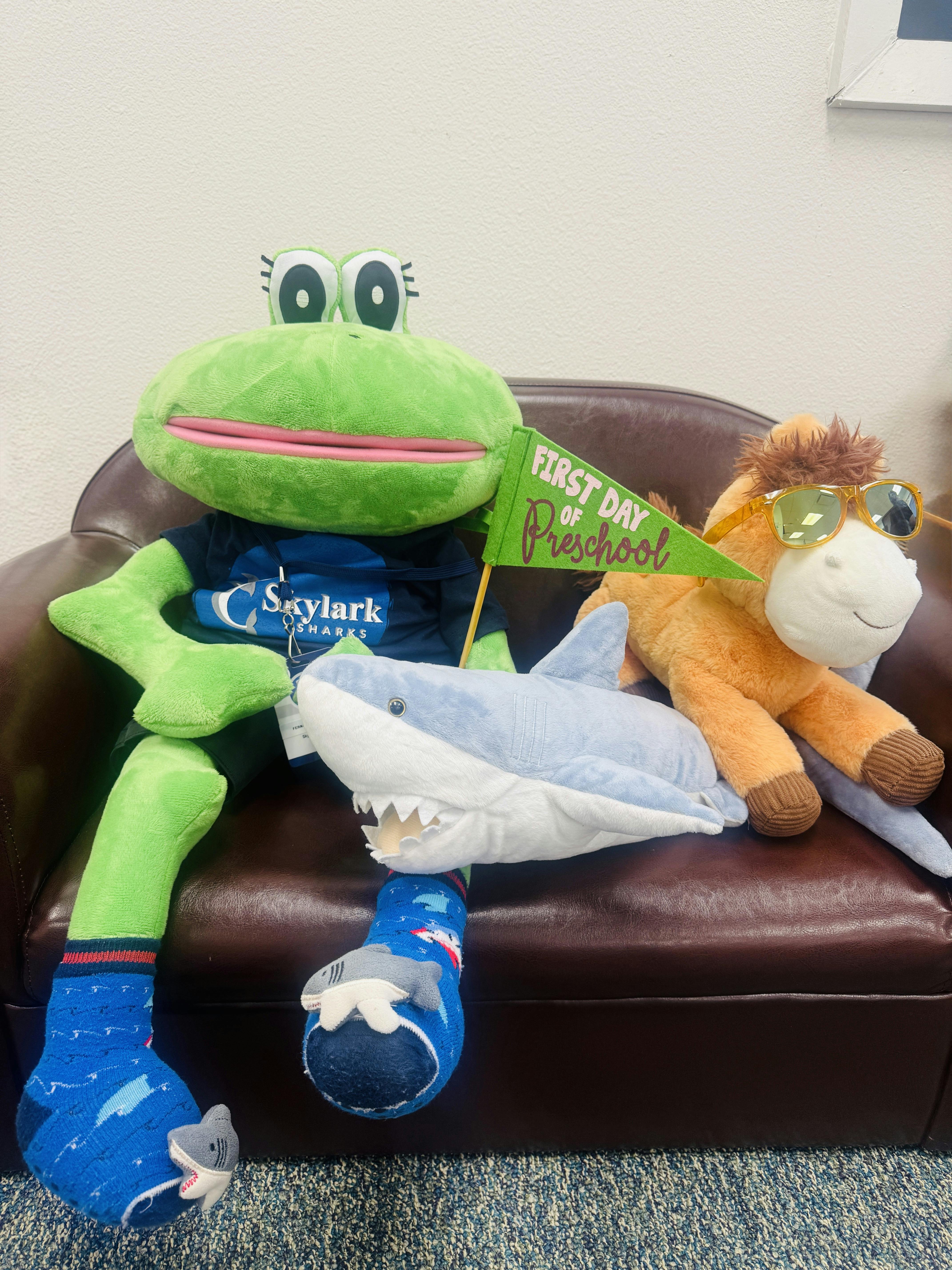 Plush frog, shark, and horse toys on a chair holding a First Day of Preschool flag