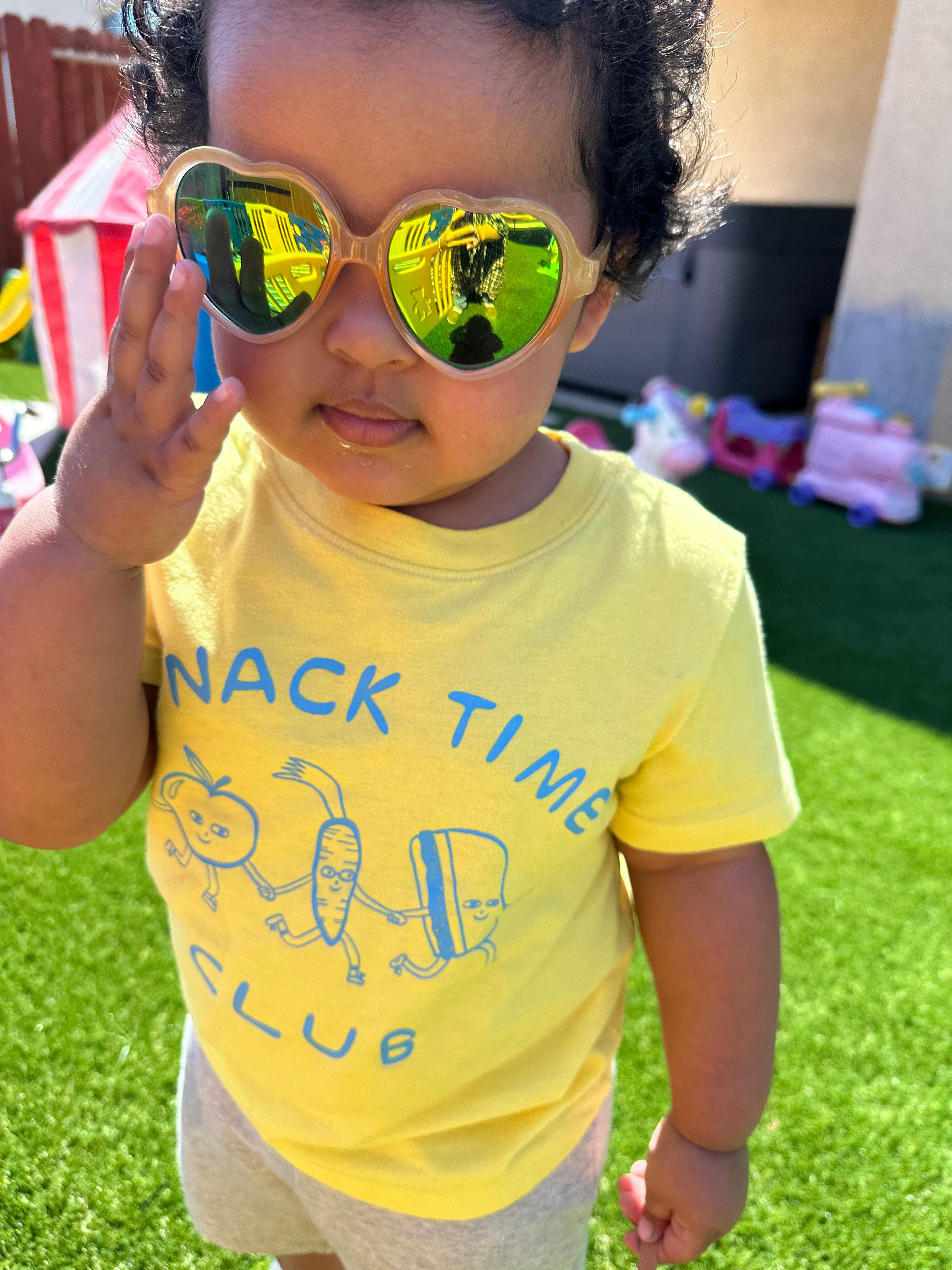 Child wearing heart-shaped sunglasses and a yellow shirt with "SNACK TIME CLUB"
