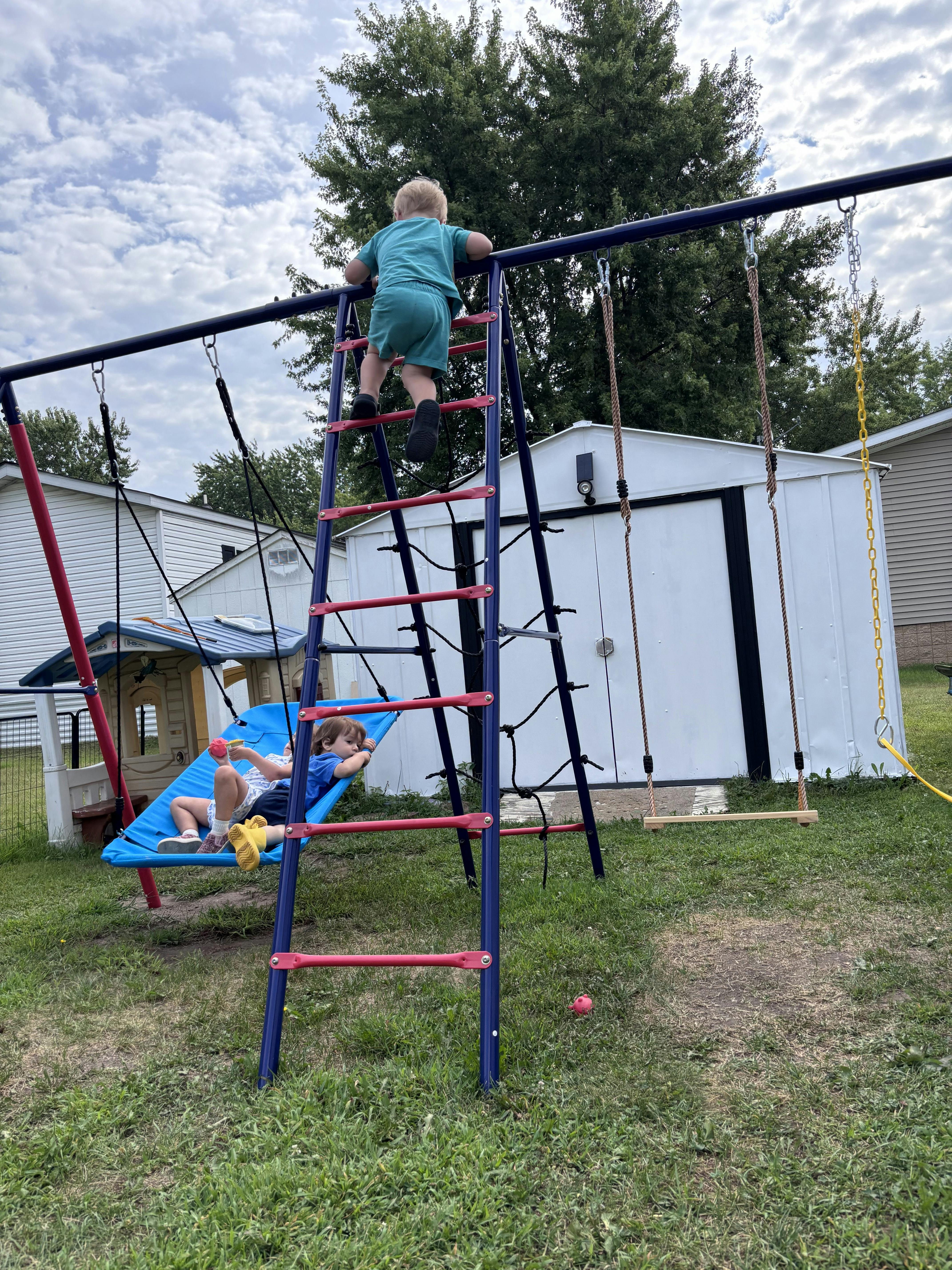 Child climbing a playset ladder while another child relaxes in a swing