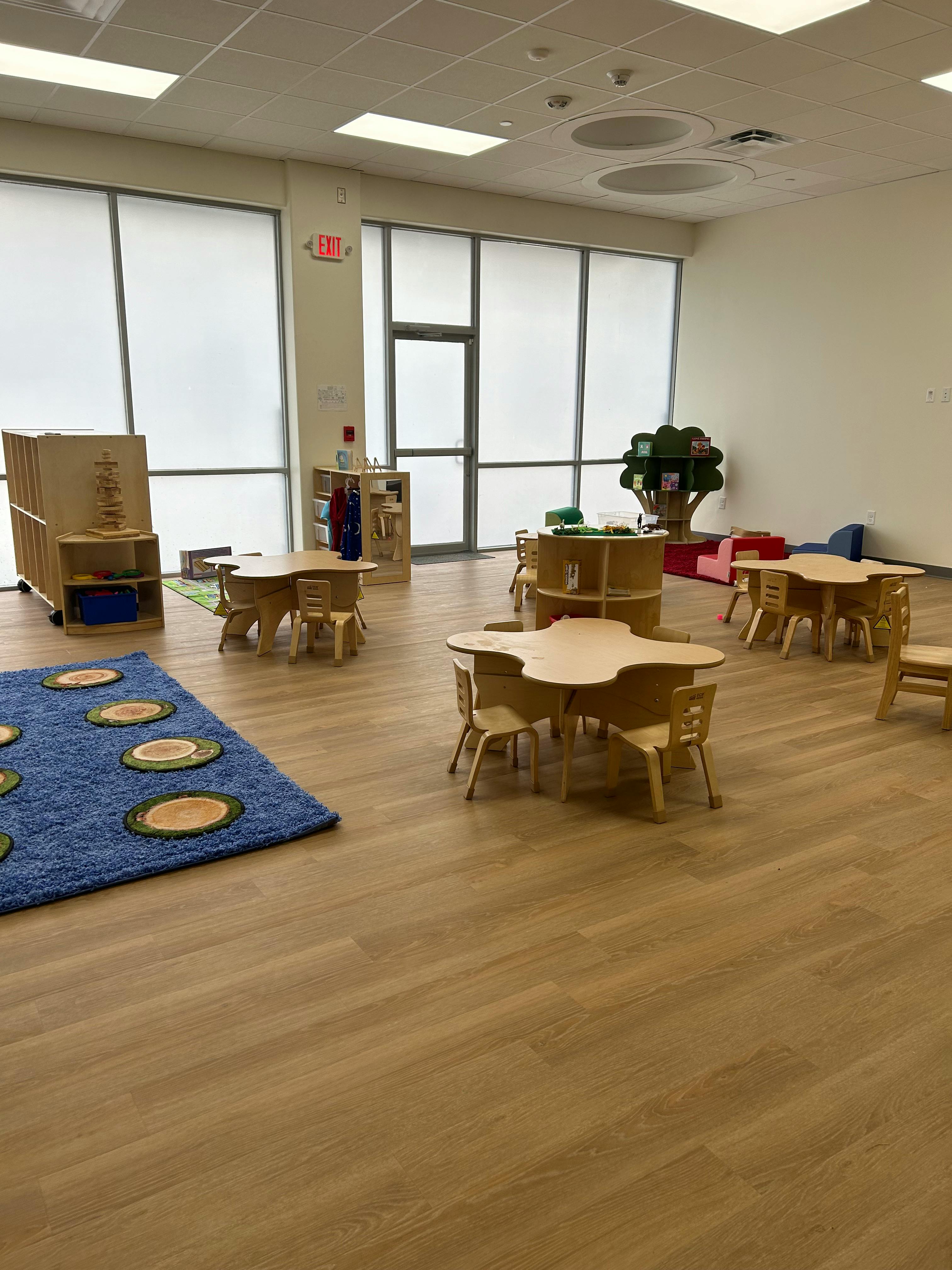 Empty classroom with small tables and chairs, a blue rug, and large windows