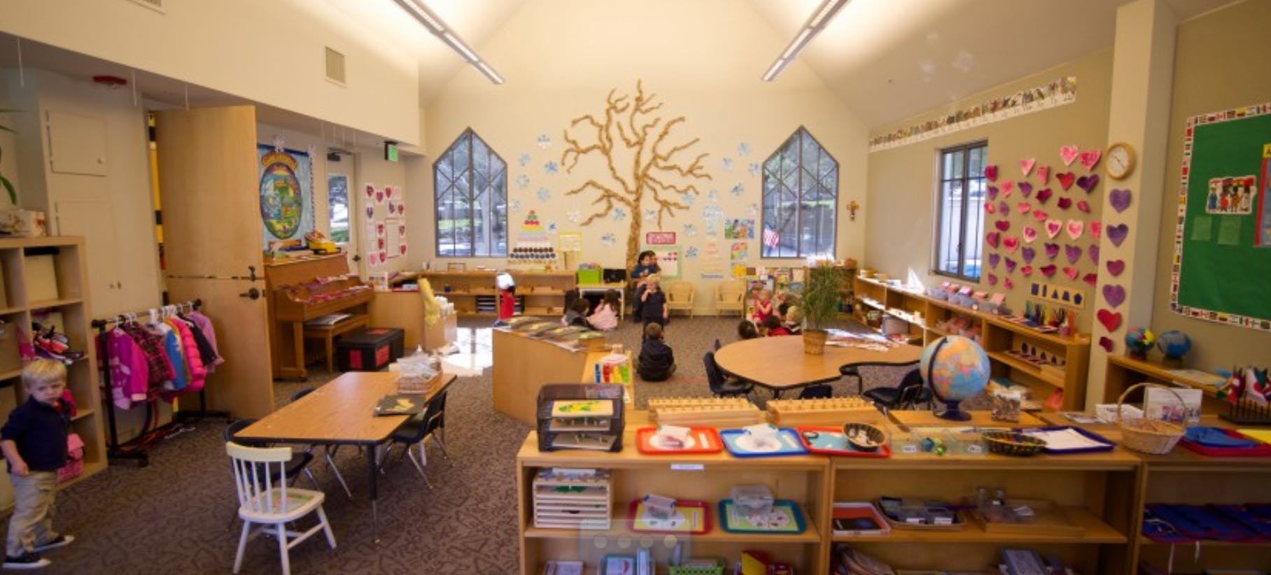 Child standing in a classroom with tables, chairs, and shelves