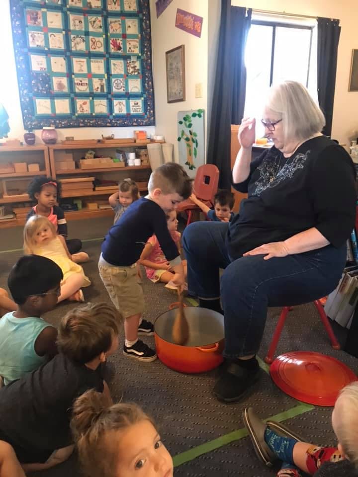 Adult and children gathered around a child stirring a pot with a wooden spoon