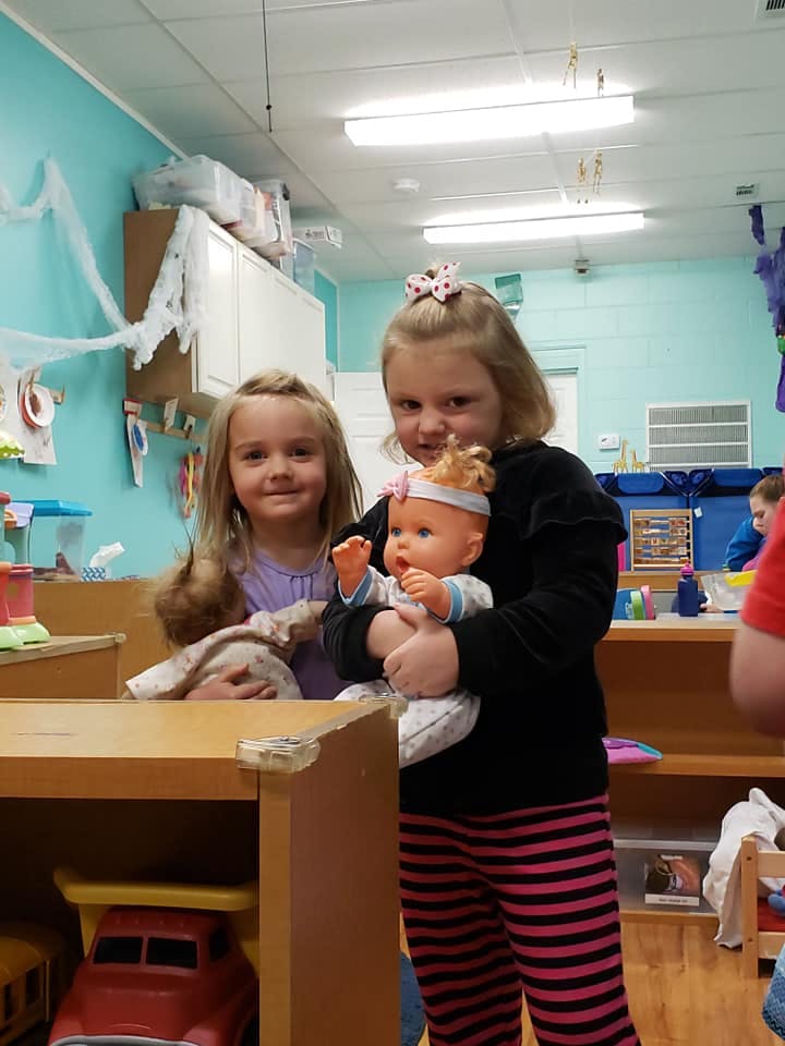 Two children in a classroom, one holding a doll