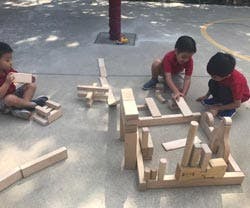 Three children playing with wooden blocks on a paved surface