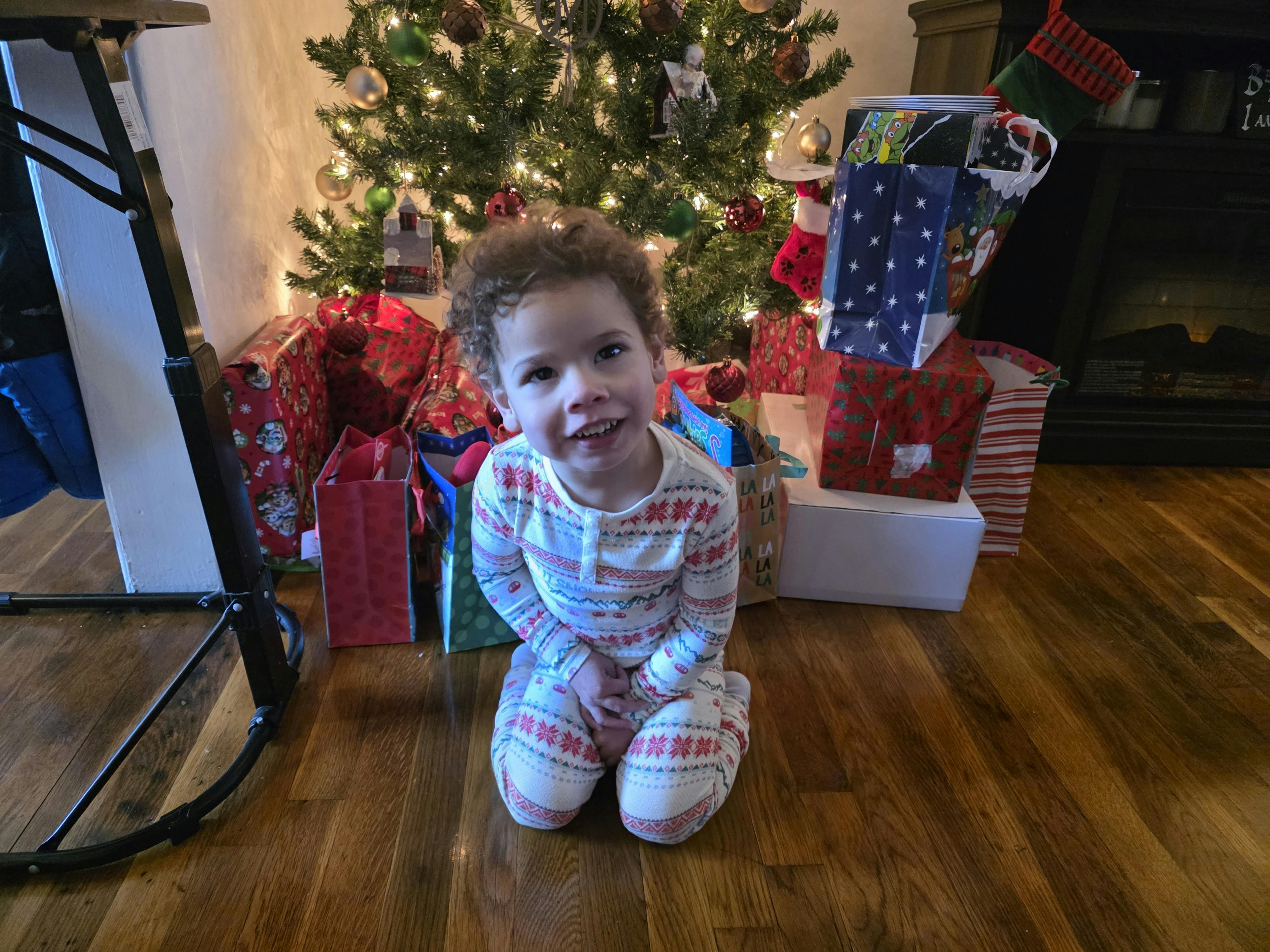 Child in pajamas kneeling on the floor in front of a Christmas tree and gifts