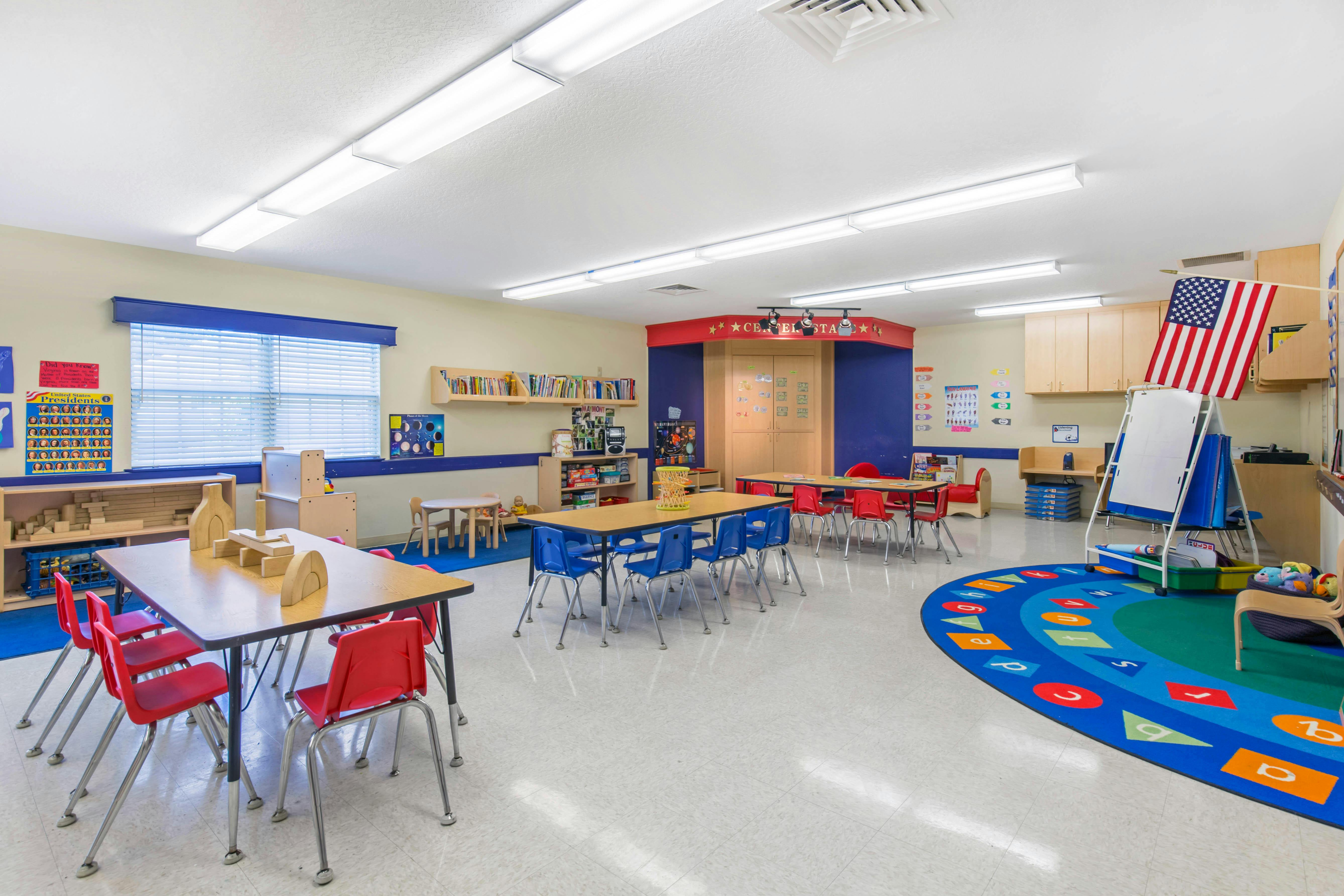 Empty classroom with small tables and chairs, a rug with letters, and an American flag