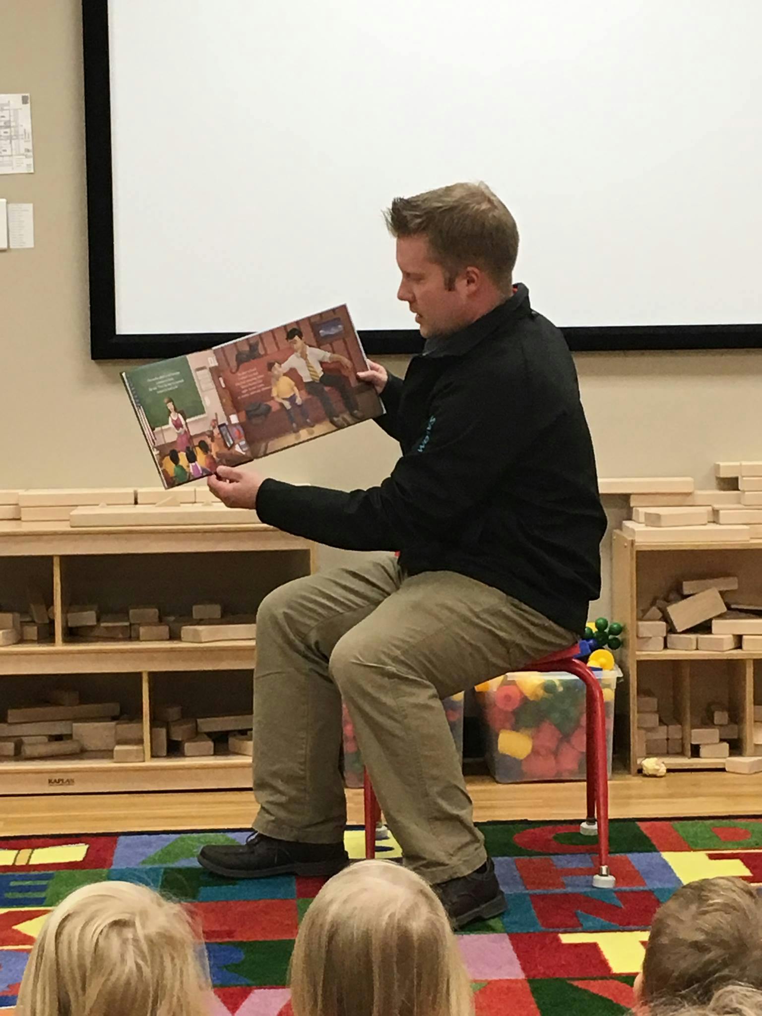 Person sitting on a stool reading a book to children in a classroom
