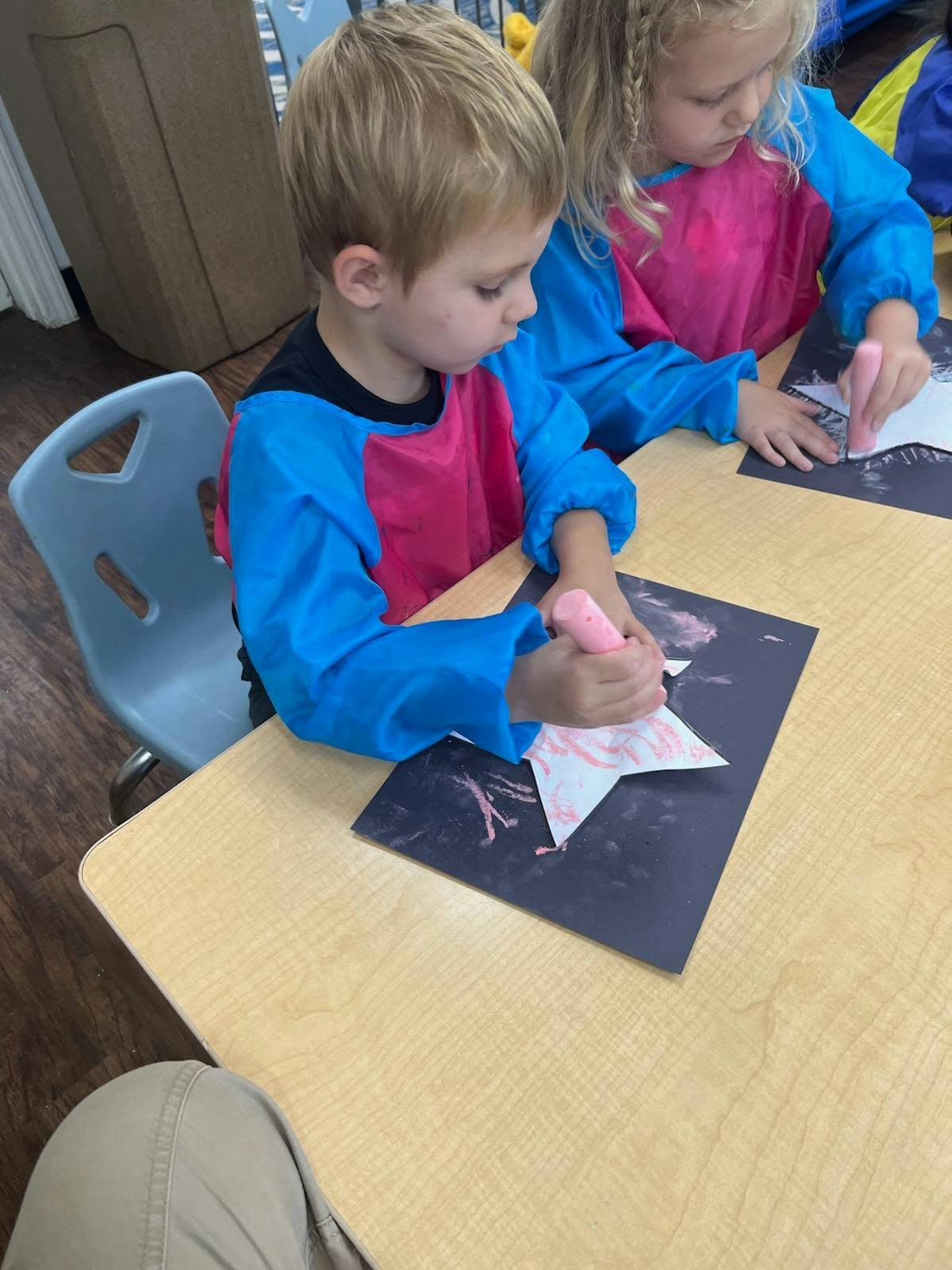 Two children coloring star cutouts with chalk at a table