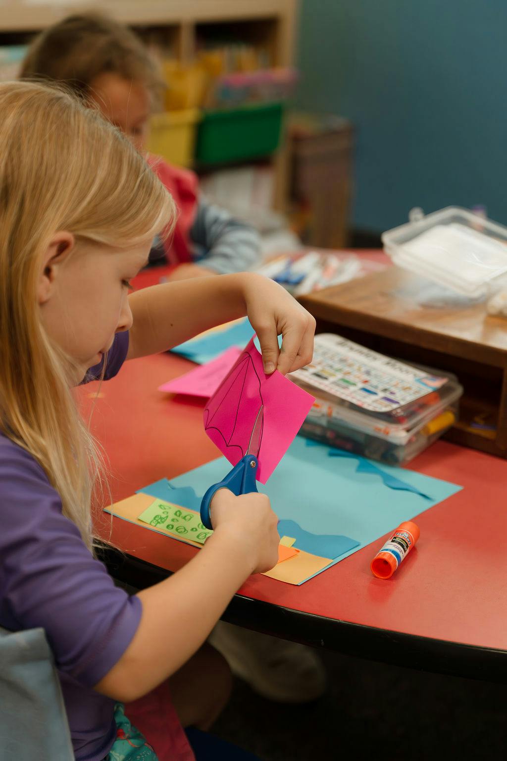 Child cutting pink paper with scissors at a classroom table