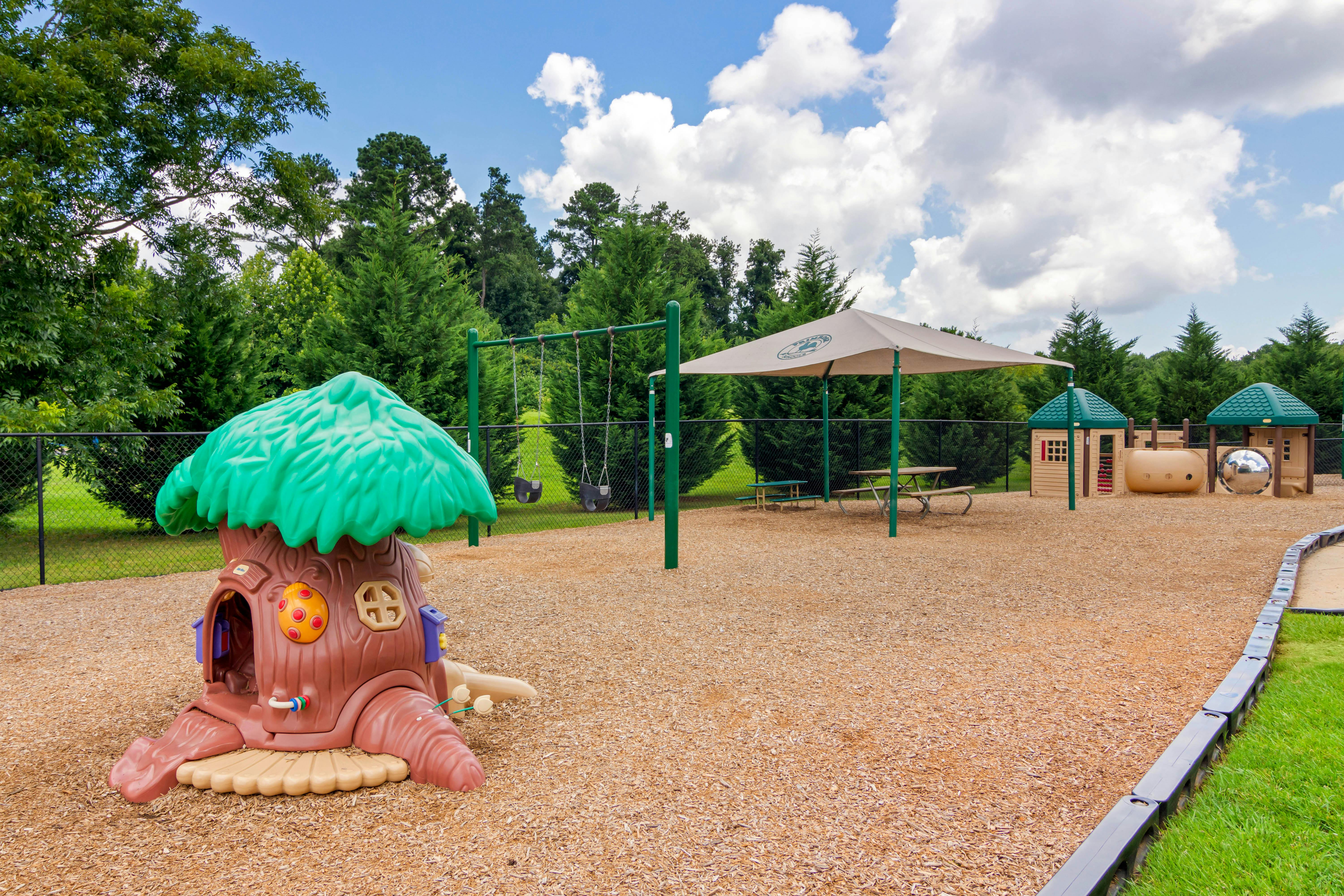 A fenced playground with wood chips, swings, playhouses, and an umbrella