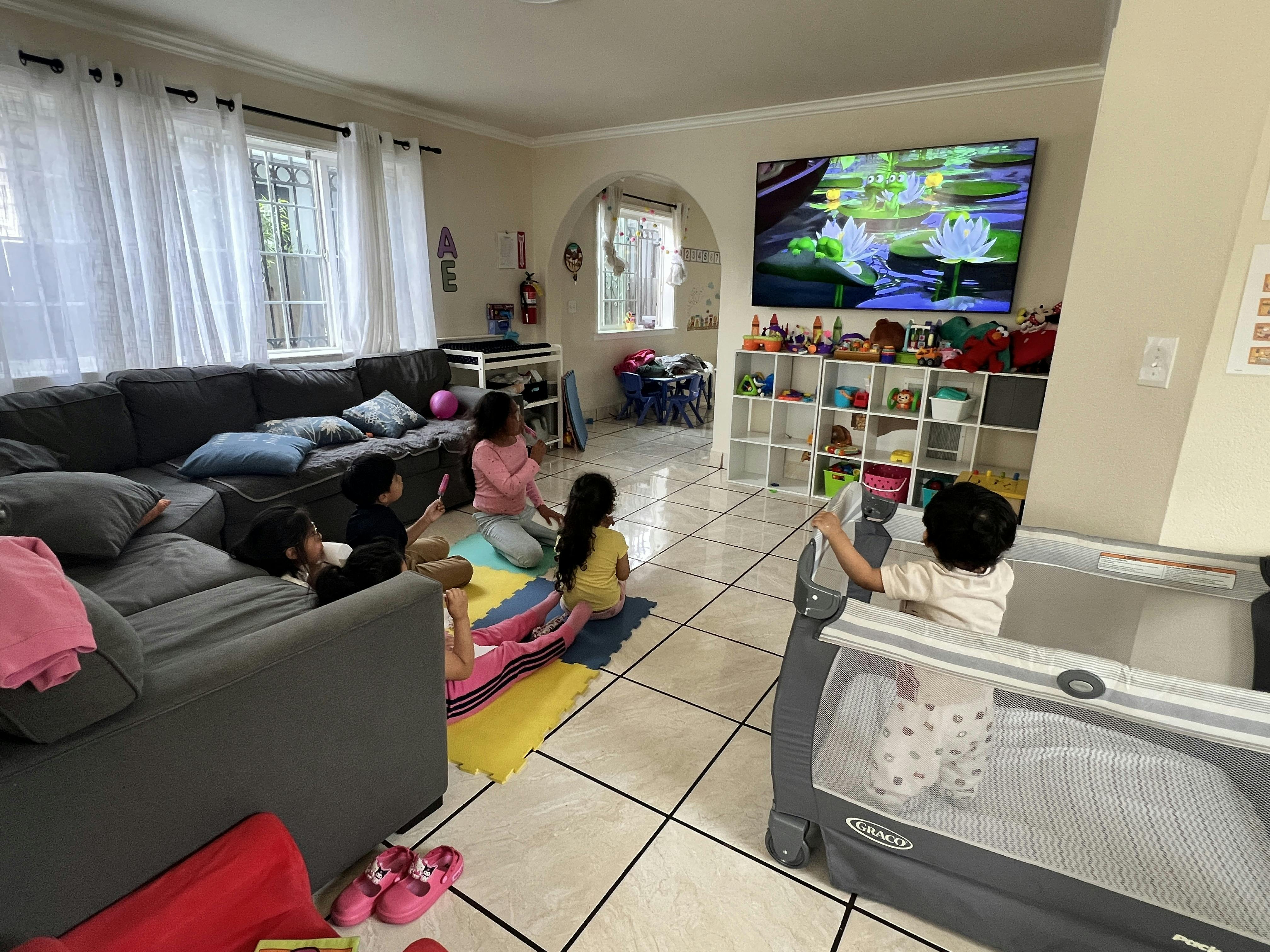 Children sitting on floor mats and a child in a playpen watching a television