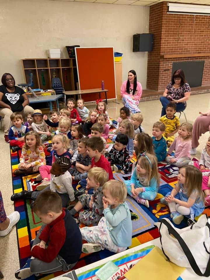 Many children sitting on a colorful rug with three adults in a classroom