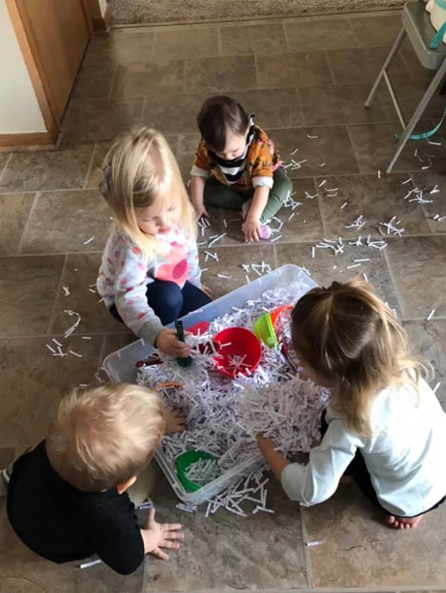 Four children playing with shredded paper in a container on the floor