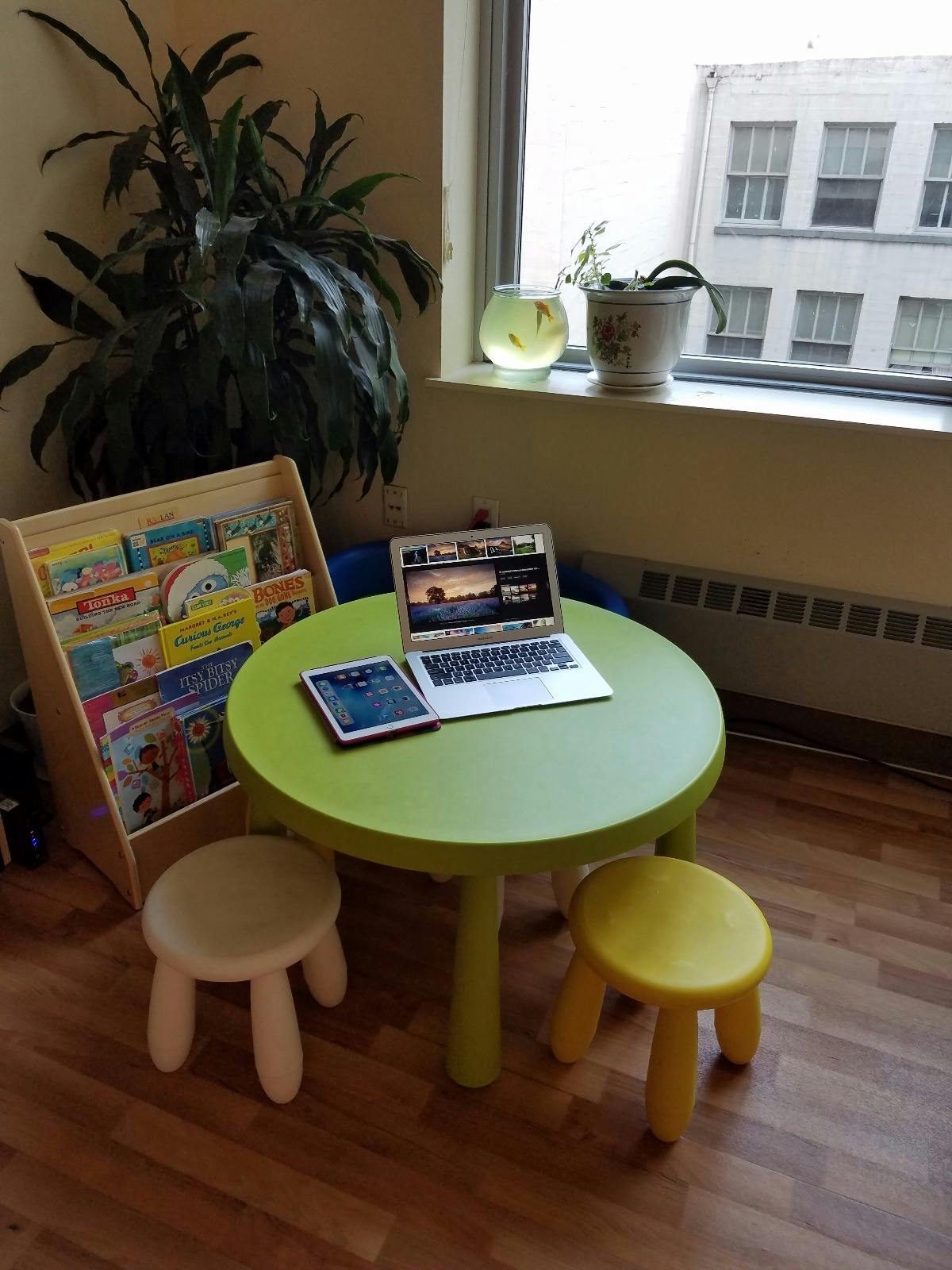 Laptop and tablet on a small green table with two stools and a bookshelf