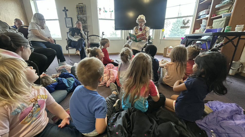 Children sitting on the floor watching an adult in a white bonnet read a book
