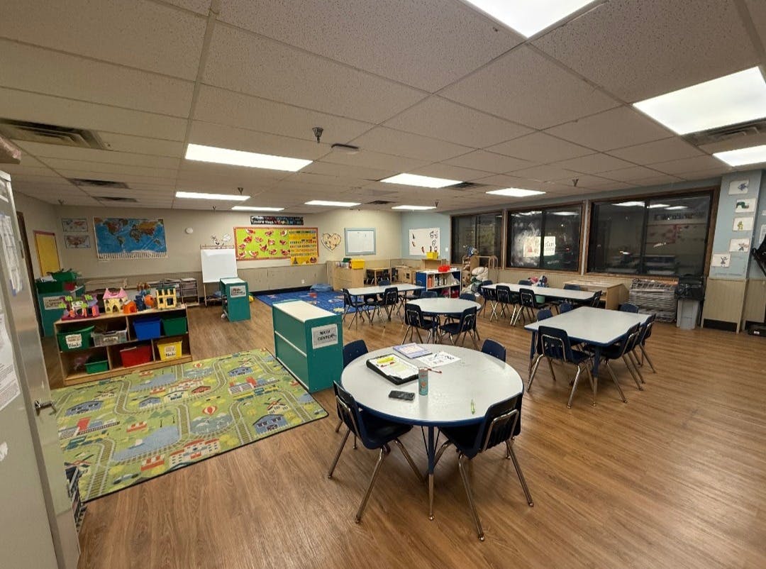 Empty classroom with small tables and chairs, a play rug, and shelves with toys