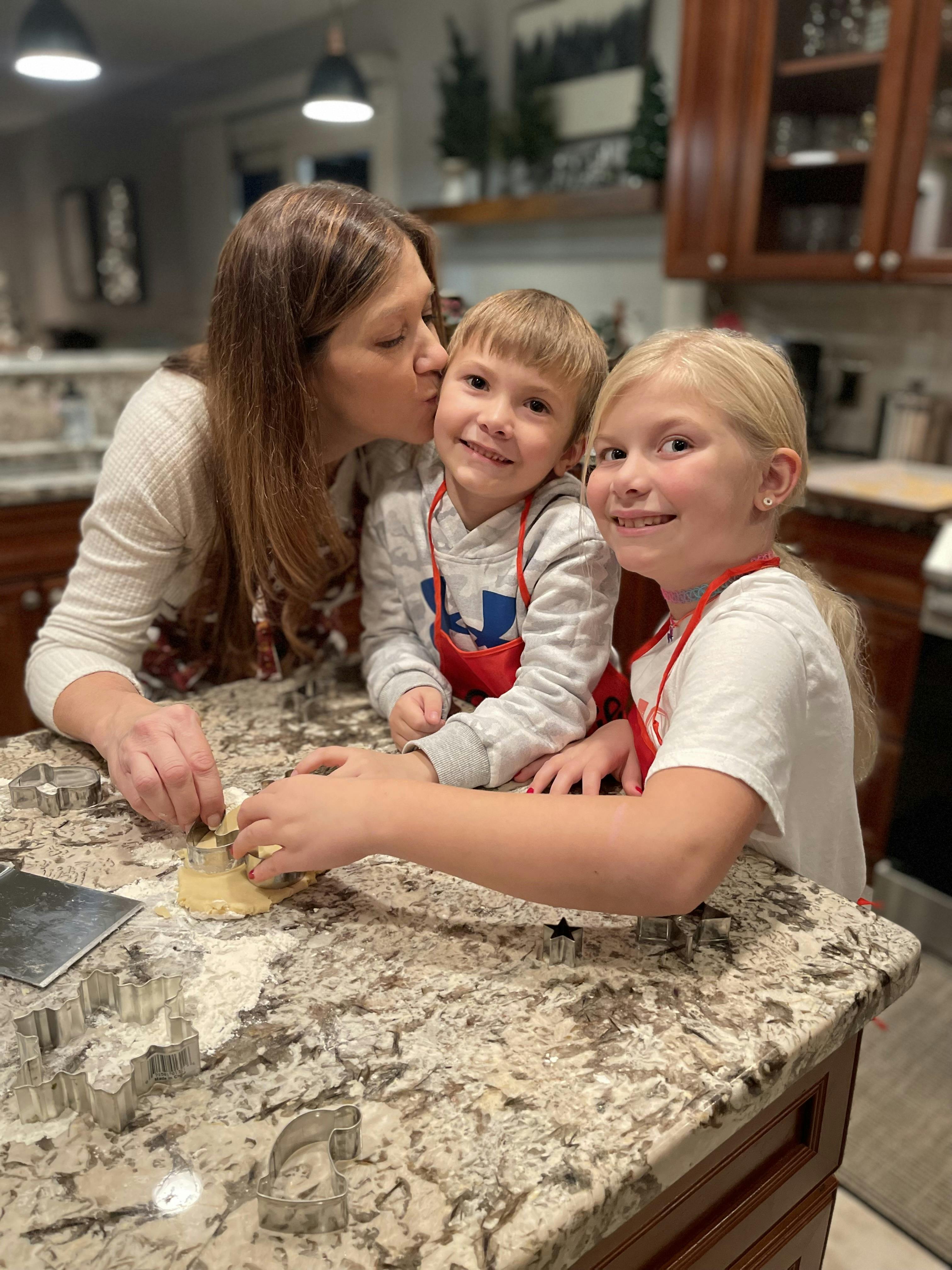 A person kissing a child while another child cuts cookie dough in a kitchen