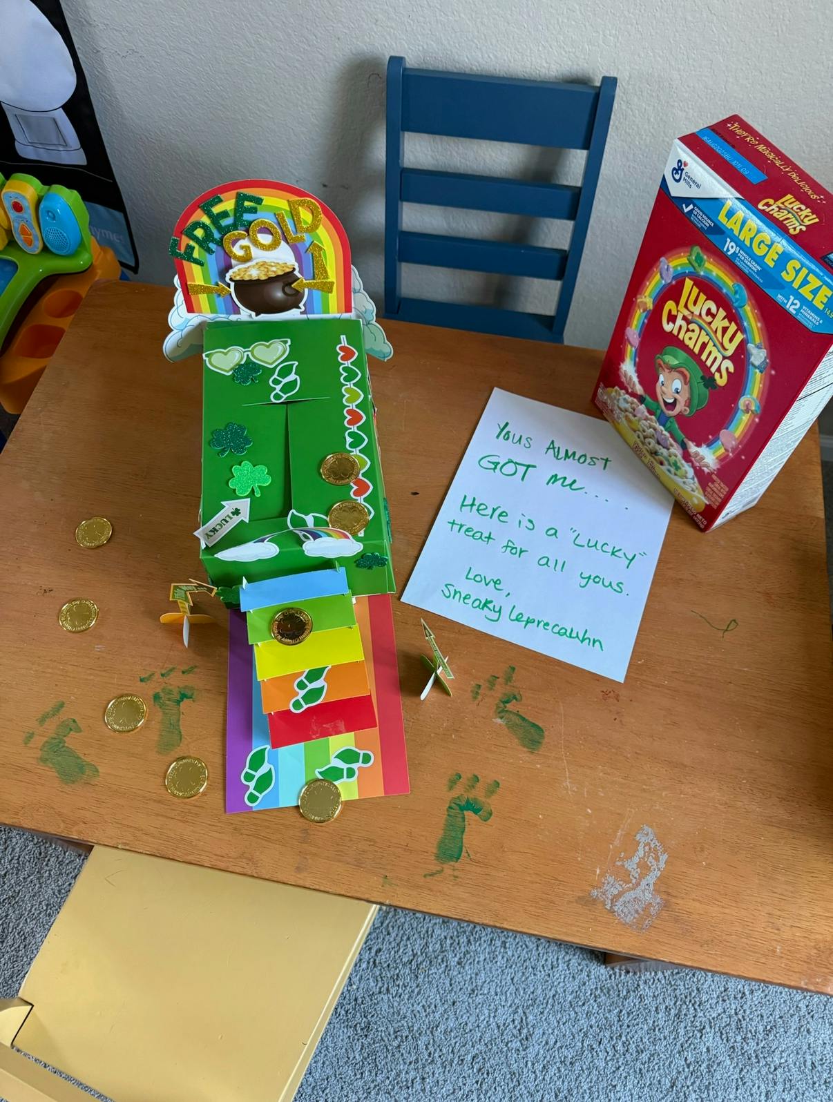 Lucky Charms box, green craft, and note on a wooden surface with gold coins and green footprints