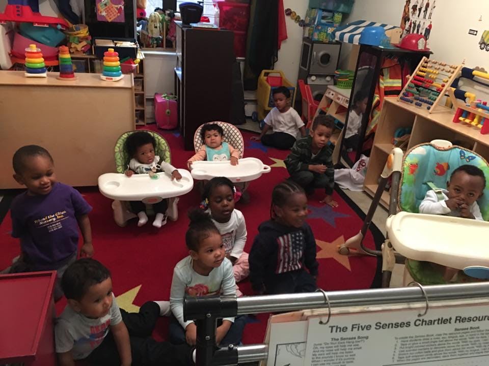 Multiple children in a classroom, some in high chairs, some on the floor
